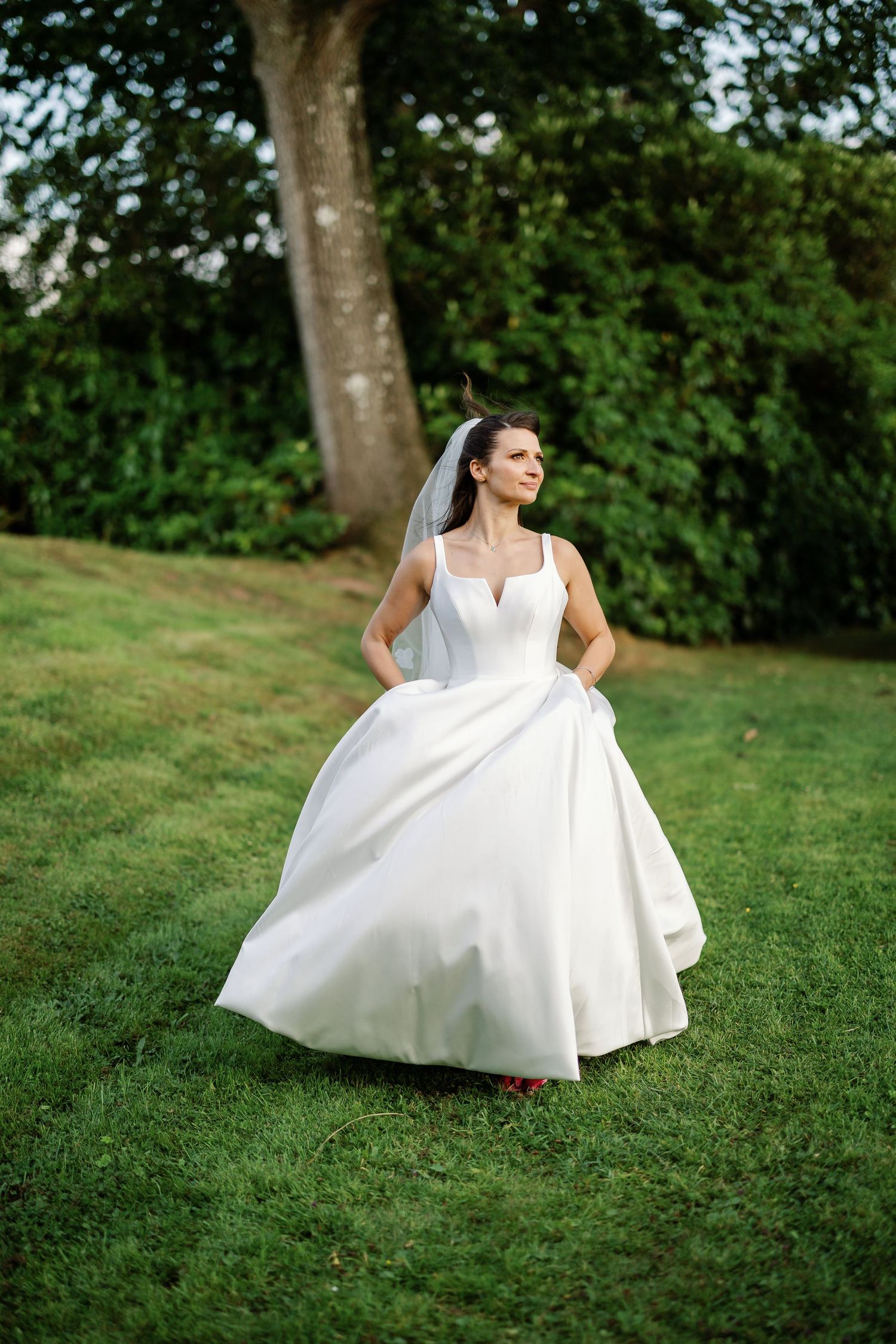 A bride in a flowing white wedding dress twirls gracefully on a grassy hillside surrounded by lush green trees.
