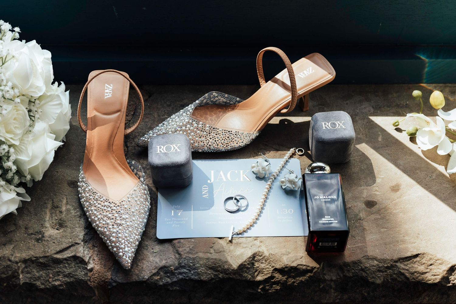 Elegant crystal-embellished wedding heels and accessories arranged with white flowers against a dark background.