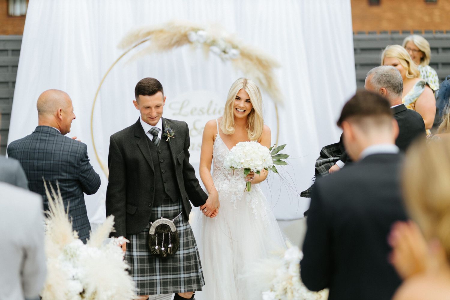 A wedding ceremony takes place under a white arch decorated with dried pampas grass and floral arrangements.