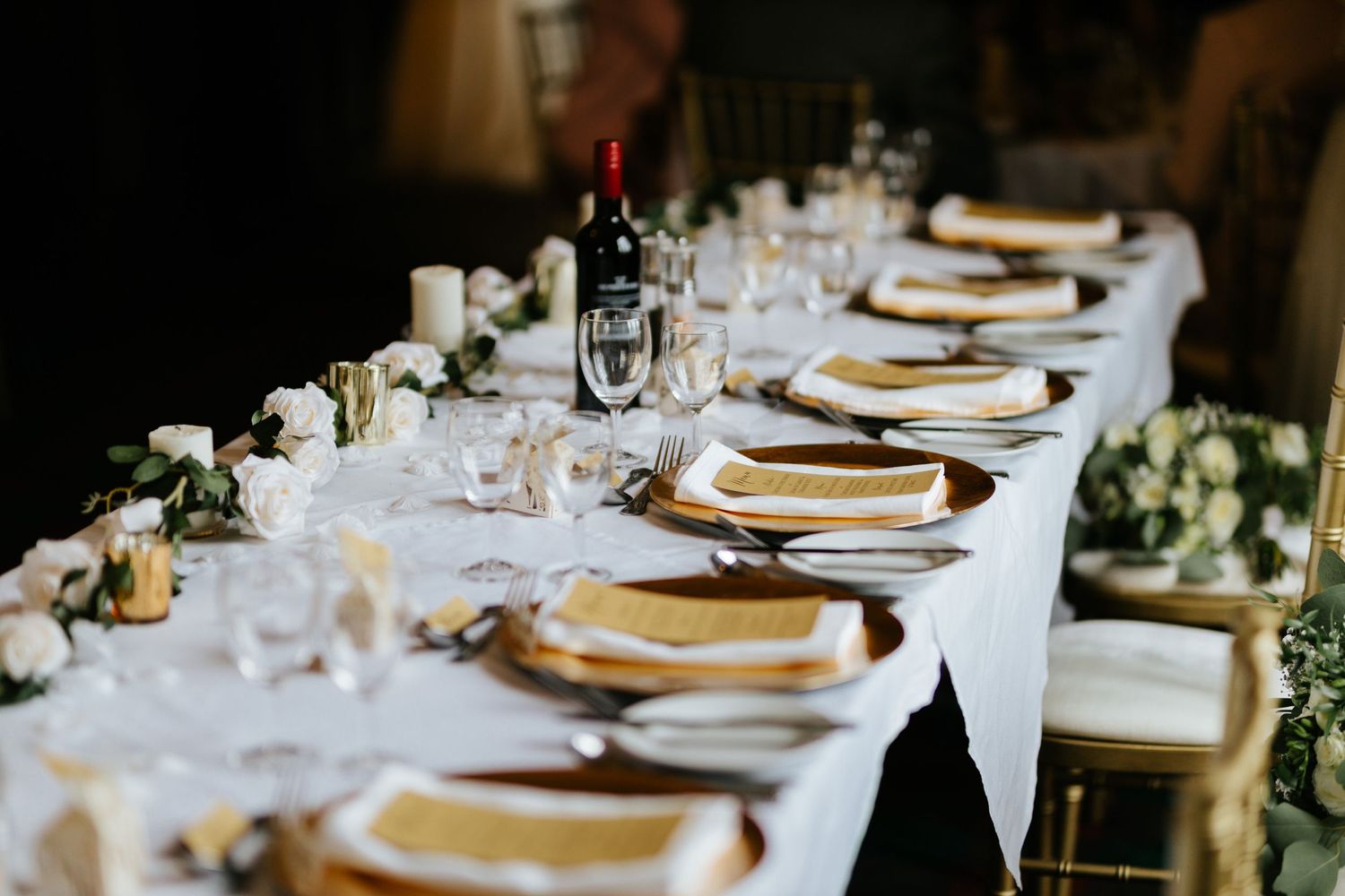 An elegant wedding table setting with gold charger plates, wine glasses, white linens, and floral centerpieces.
