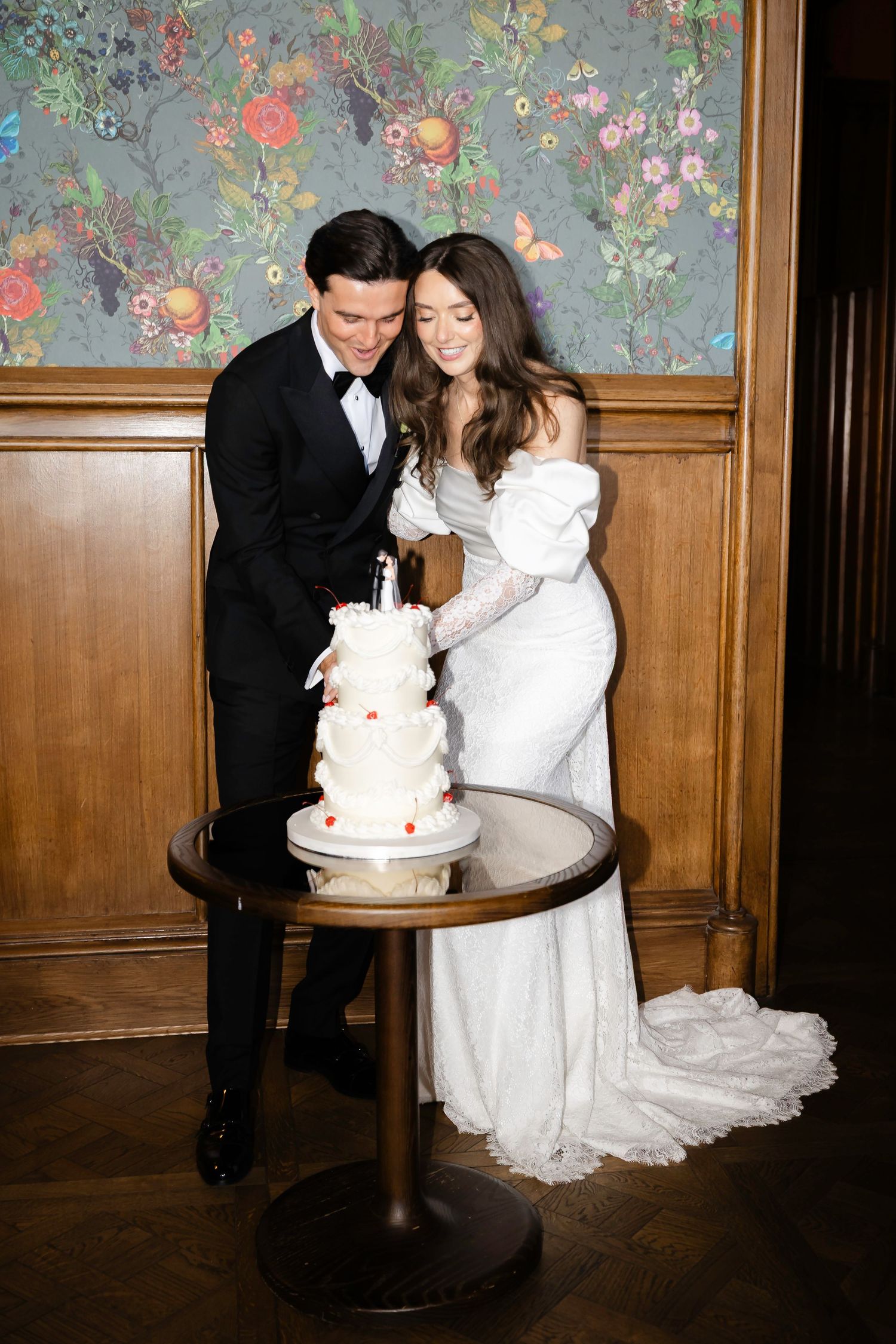 Bride and groom cutting white tiered wedding cake together in elegant wood-paneled room with floral wallpaper.