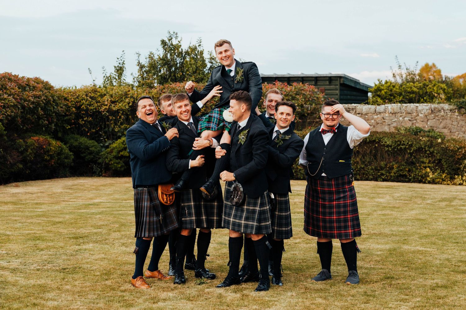 A group wearing traditional Scottish kilts and formal attire pose together on a grassy lawn during a celebration.