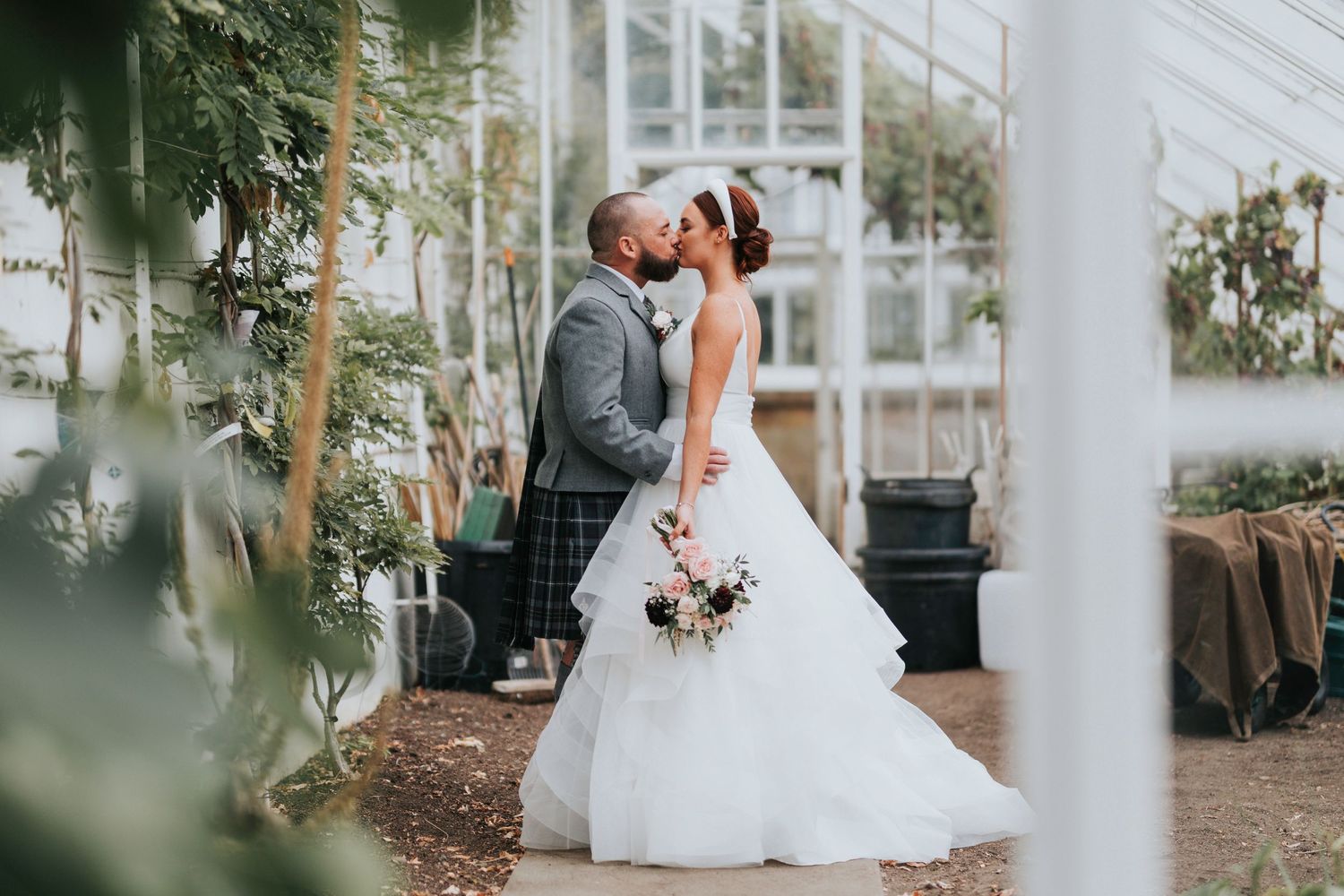 A romantic wedding couple shares a kiss in front of a greenhouse surrounded by lush greenery.