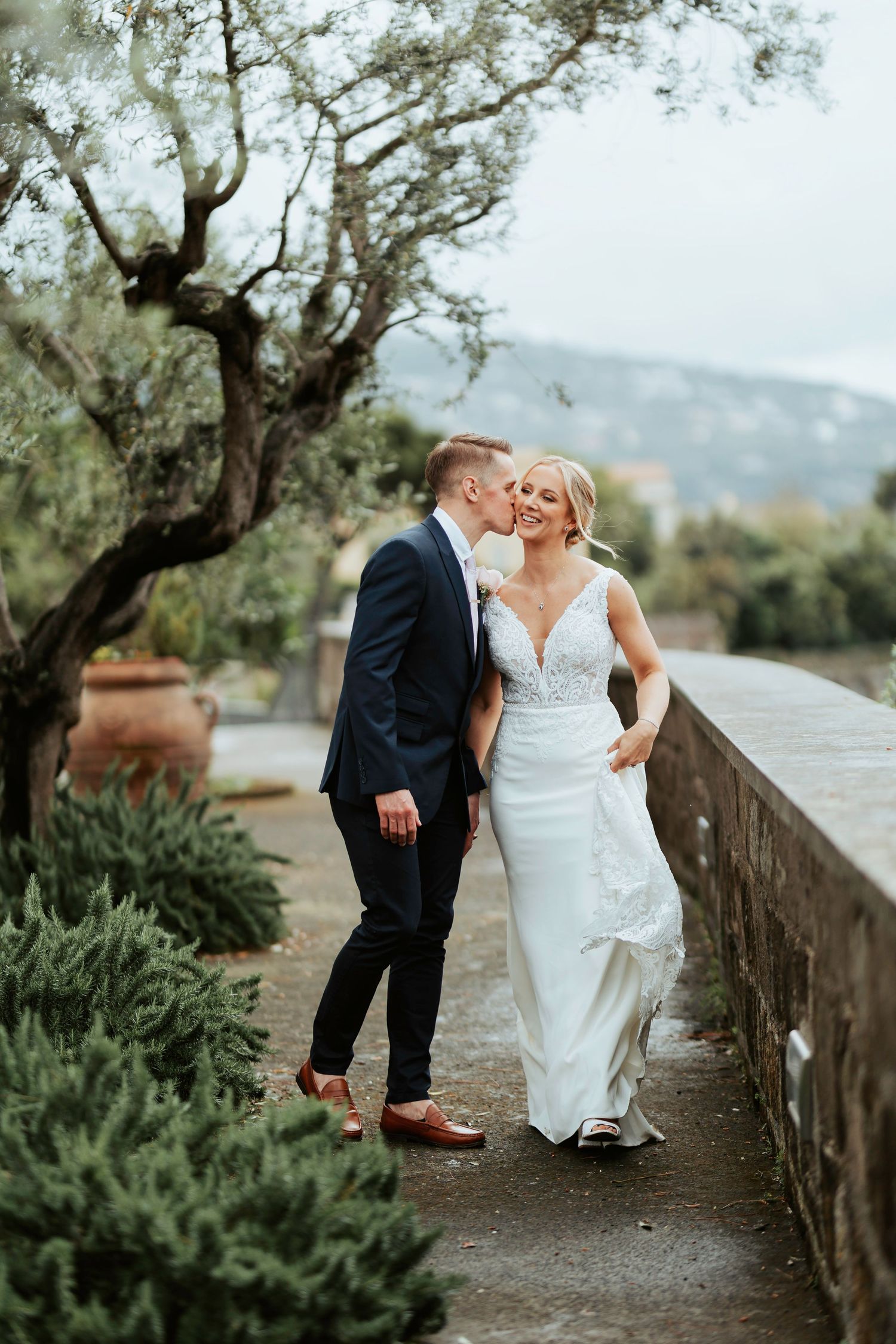 A romantic wedding couple embraces on a stone pathway surrounded by lush greenery and trees in a Mediterranean setting.