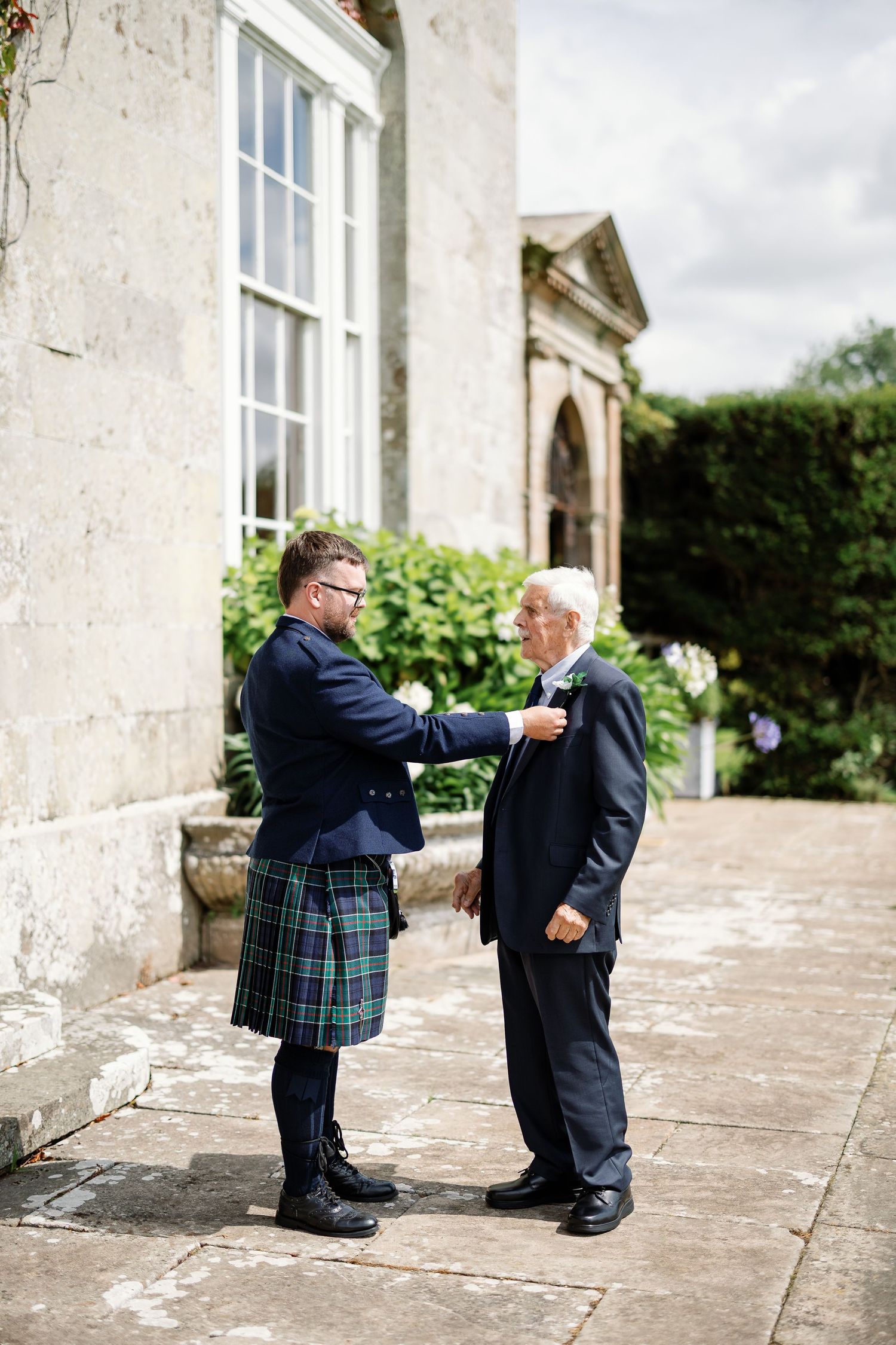Two people in formal attire with one wearing a kilt interact on a stone patio outside a historic building.
