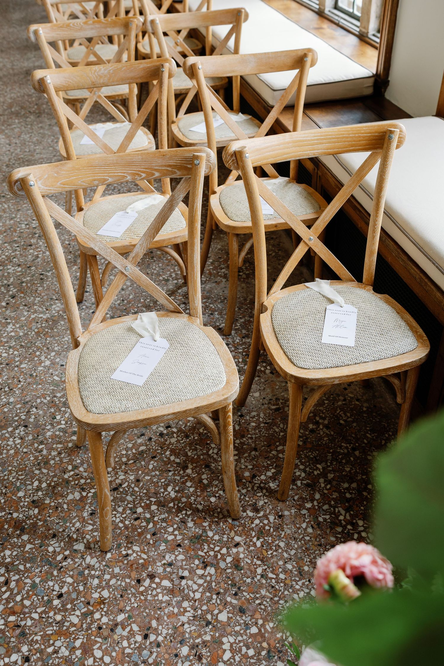 Rustic wooden crossback dining chairs with woven seats arranged in a row on a speckled floor near a window.