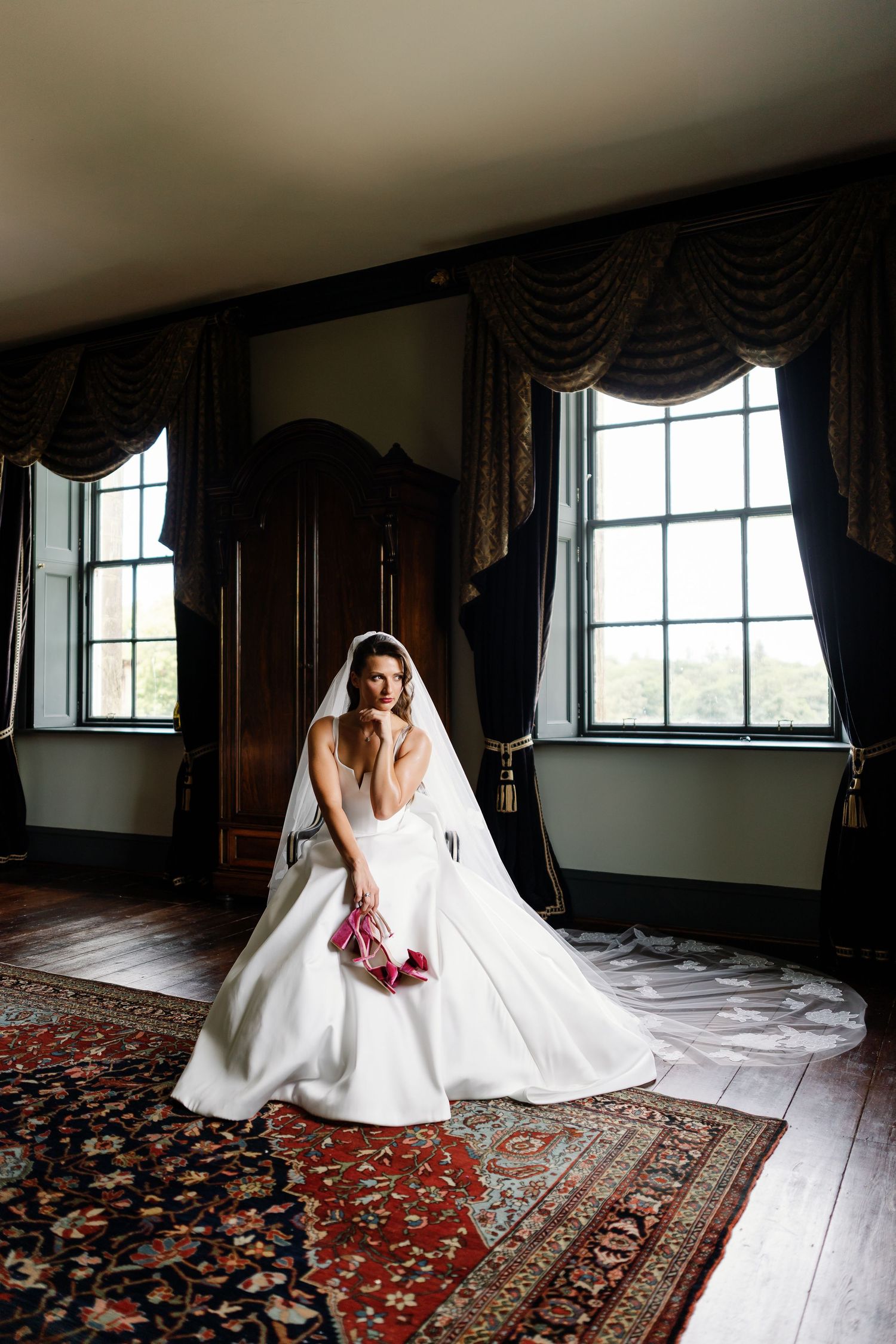 A bride in a flowing white gown sits elegantly on an ornate Persian rug in a sunlit room with large windows.
