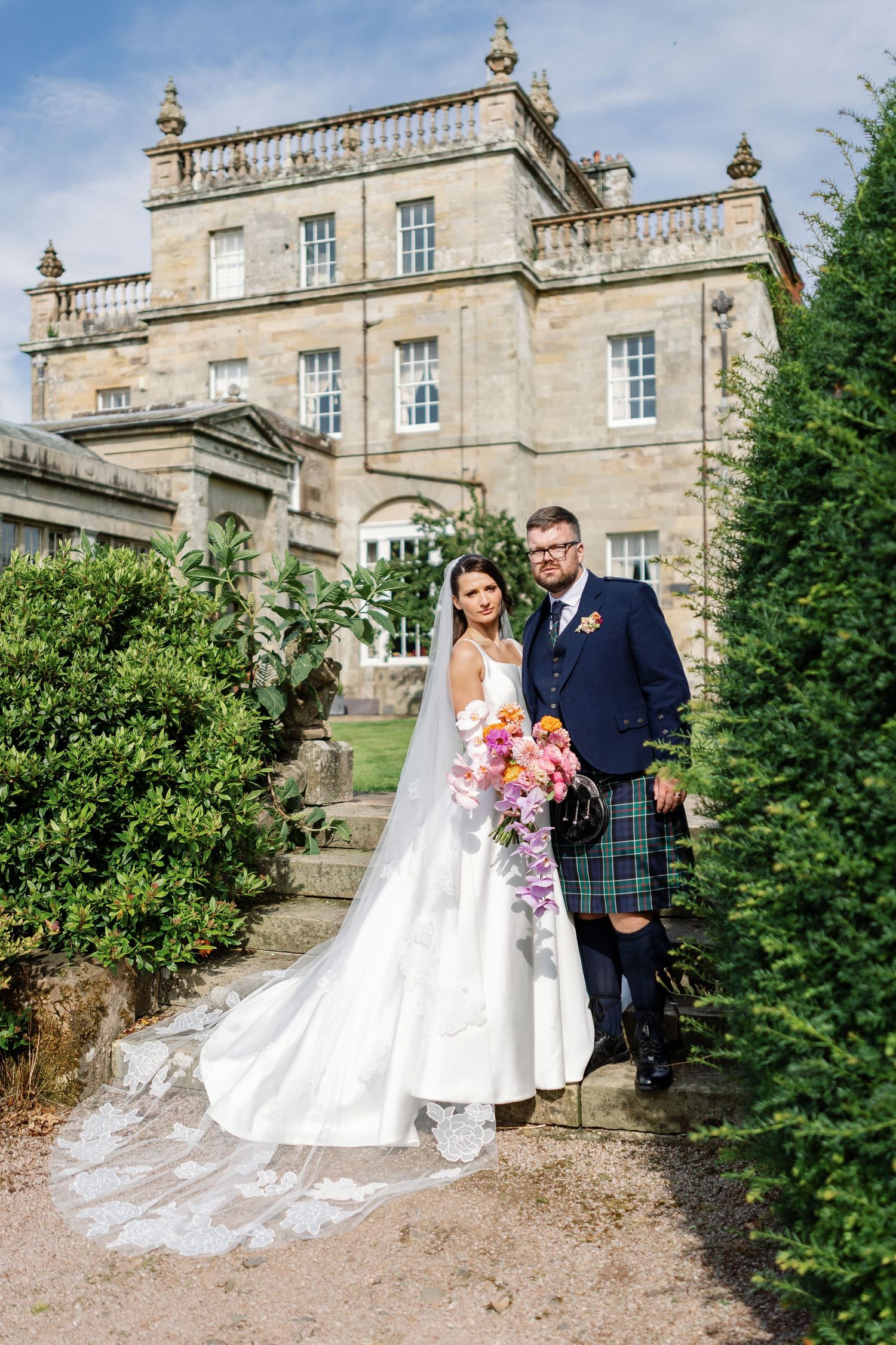 A wedding couple poses for portraits in front of a grand stone mansion with elegant architectural details on a sunny day.