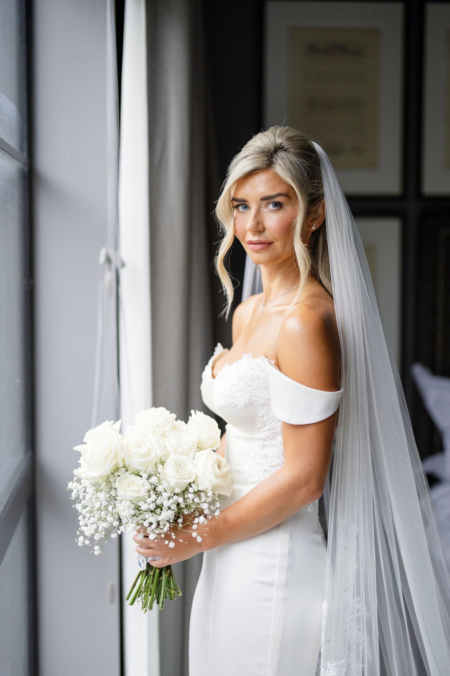 Bride in elegant white off-shoulder wedding dress holds classic white bouquet near window in natural light.