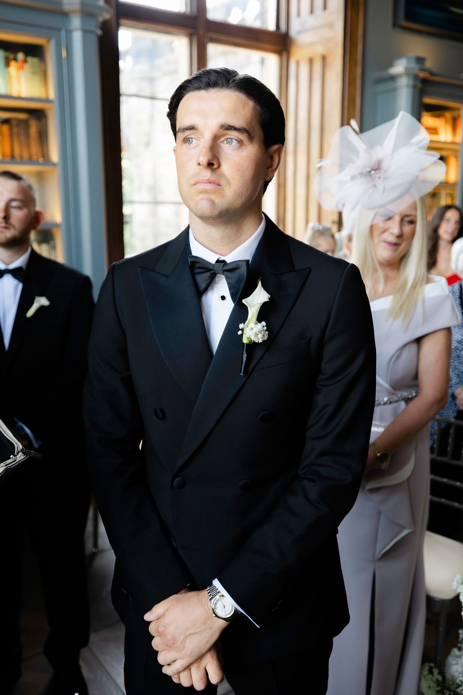 Groom in black tuxedo with white boutonniere stands at altar during wedding ceremony, guests visible in background.