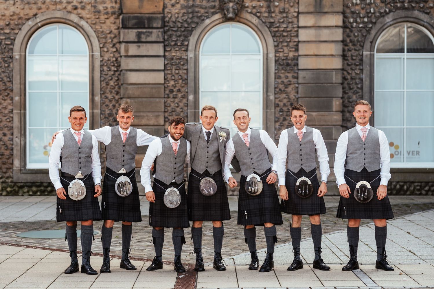 Seven groomsmen in matching grey vests and kilts pose together outside a historic stone building with arched windows.