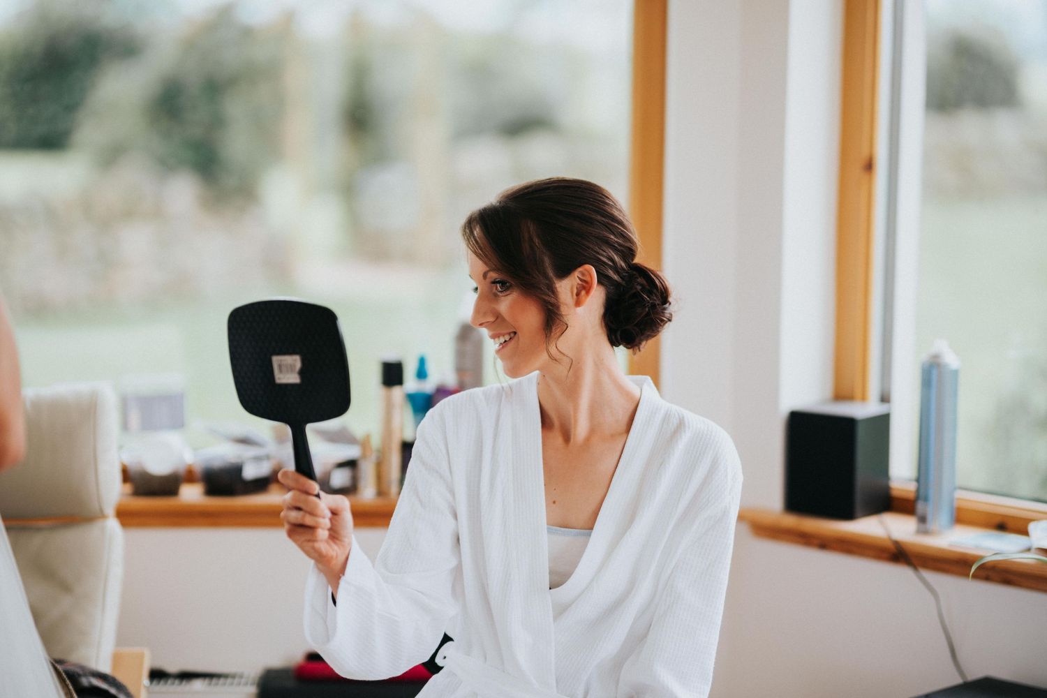 A person in a white robe holds a mirror while getting ready at a sunlit vanity table.