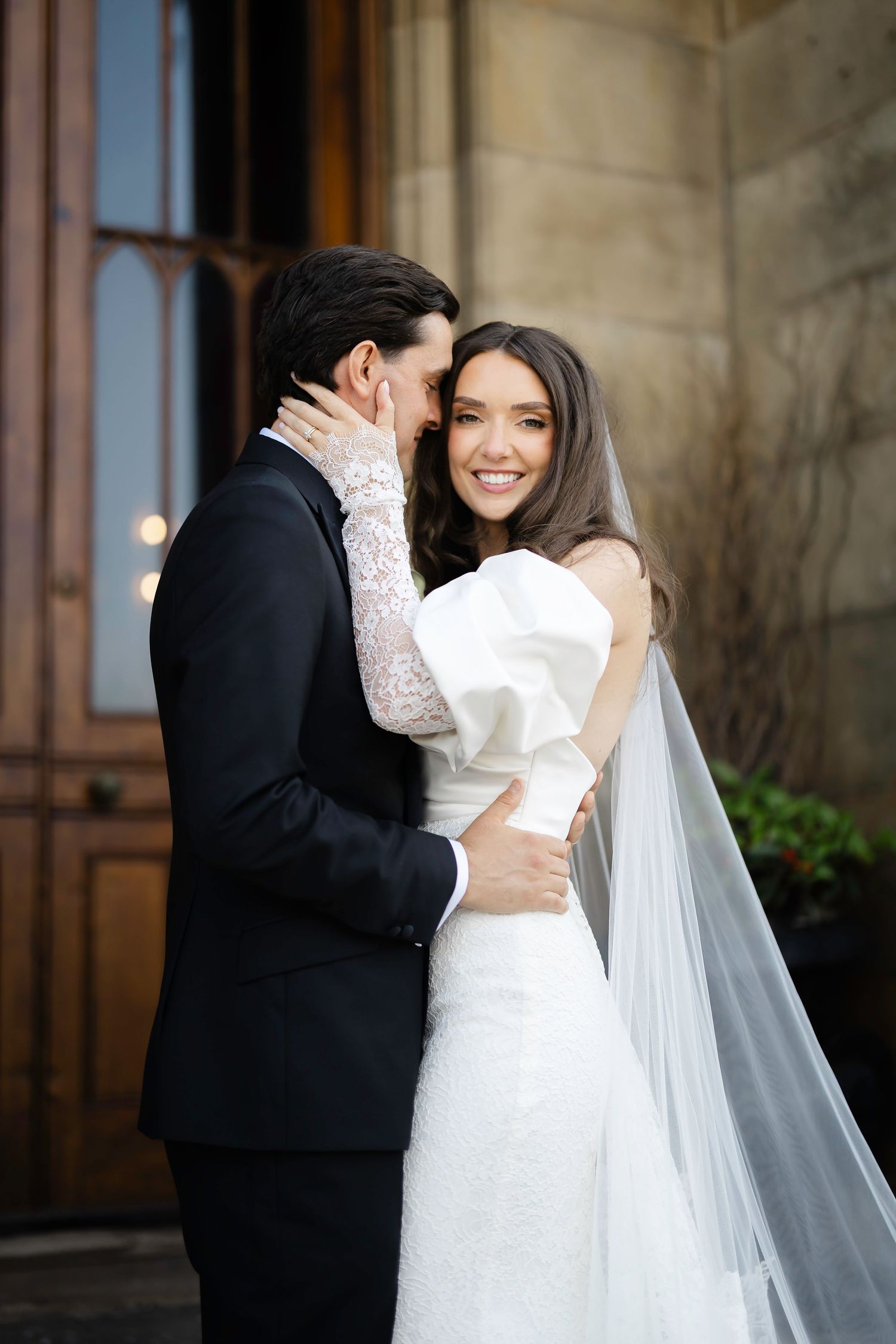 A smiling bride in a white gown and veil embraces a groom in a black suit outside a stone building.