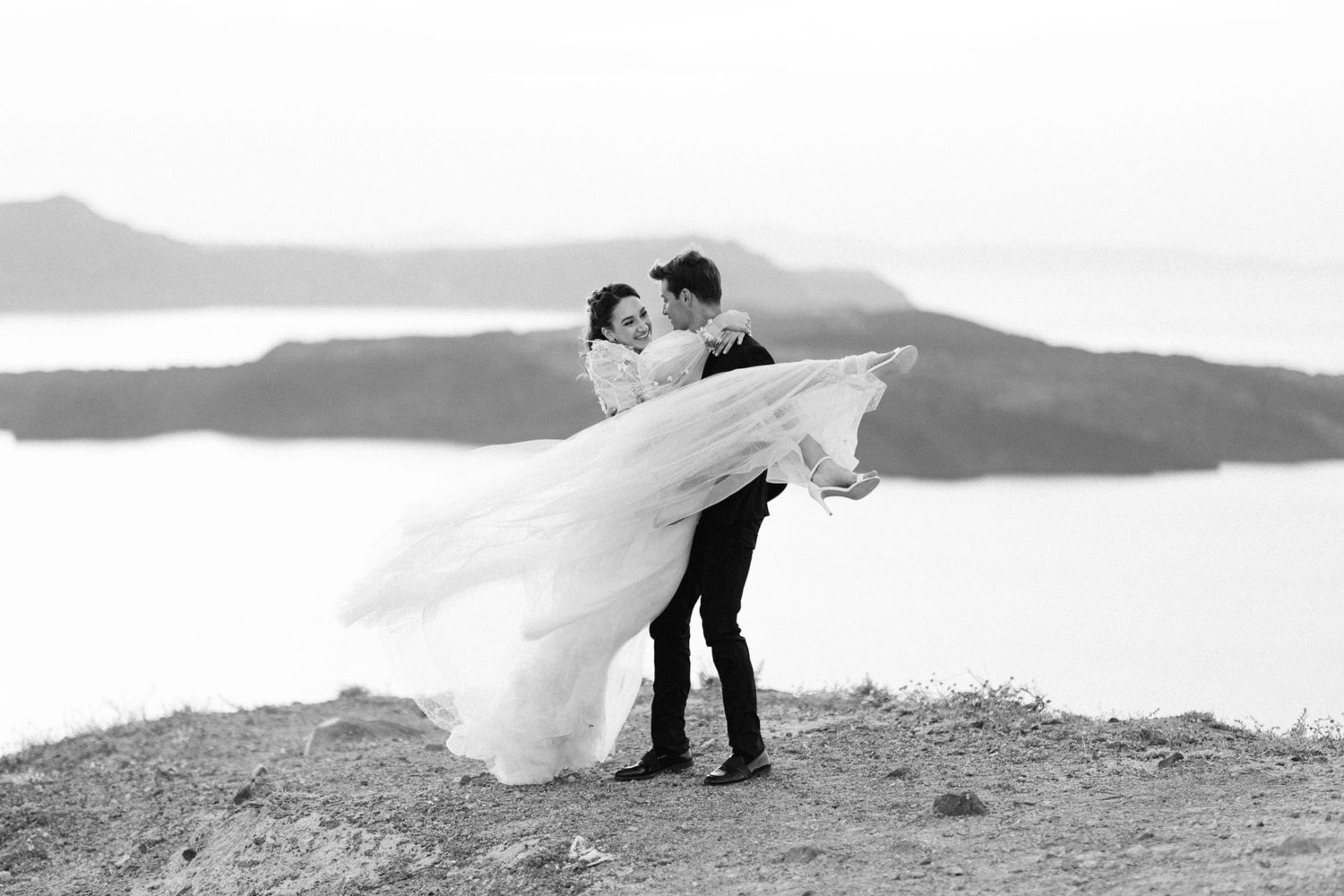 Black and white wedding photo of groom lifting bride, her gown flowing in wind, scenic coastal cliffs in background.