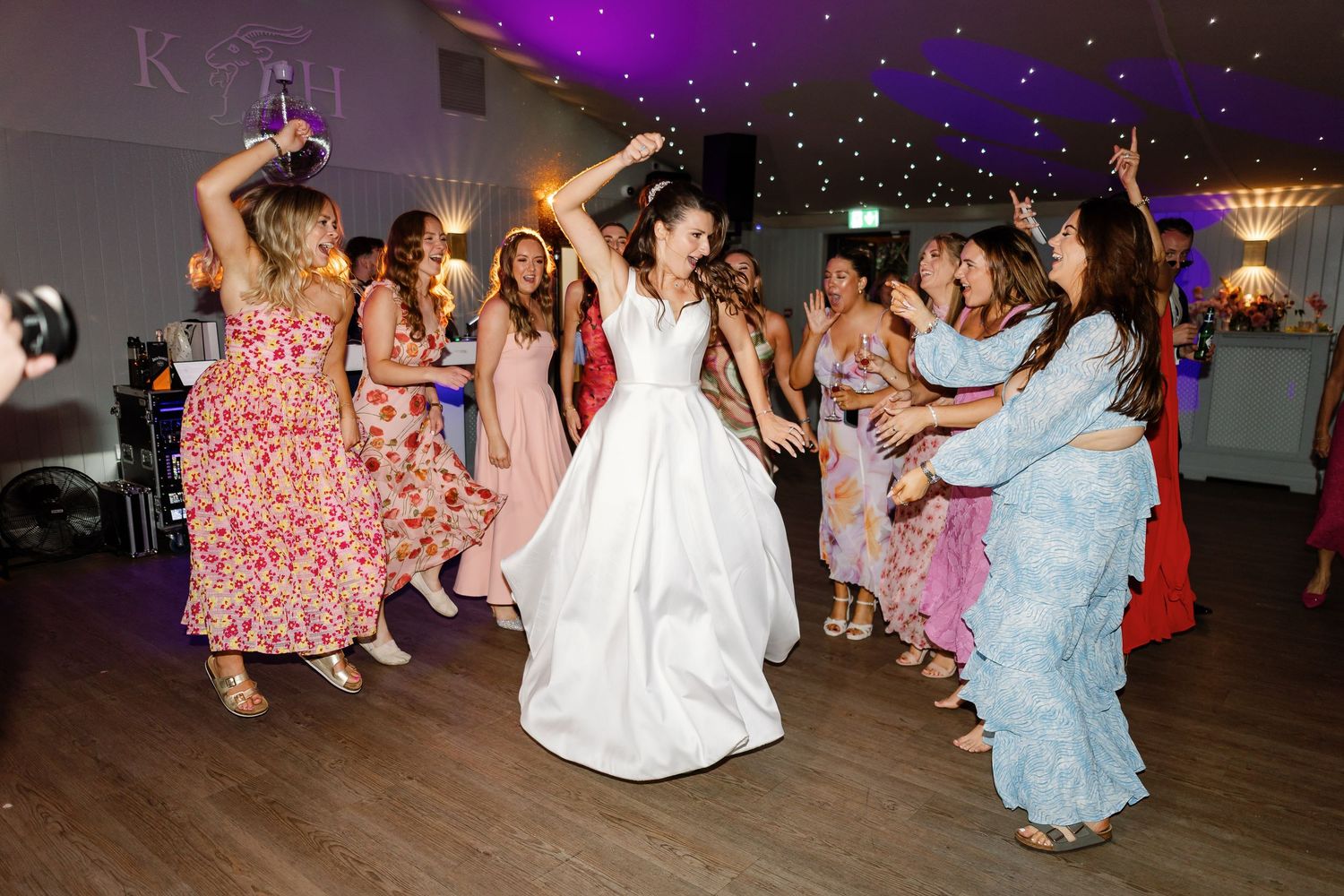 Wedding guests dance together on a dark dance floor illuminated by purple lighting at a reception celebration.