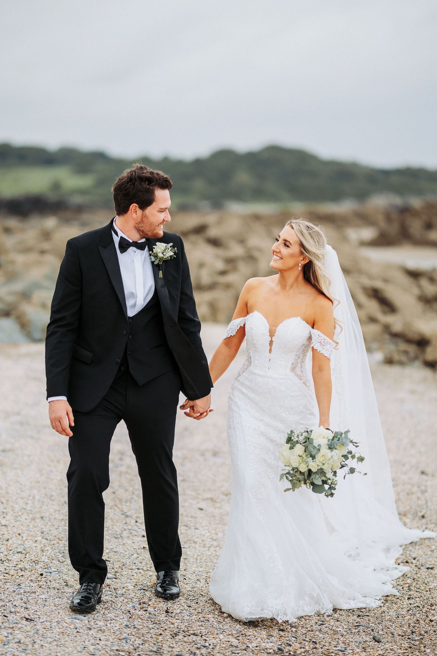 Newlywed couple walking on rocky beach path, bride in off-shoulder lace gown holding eucalyptus bouquet with ocean view.