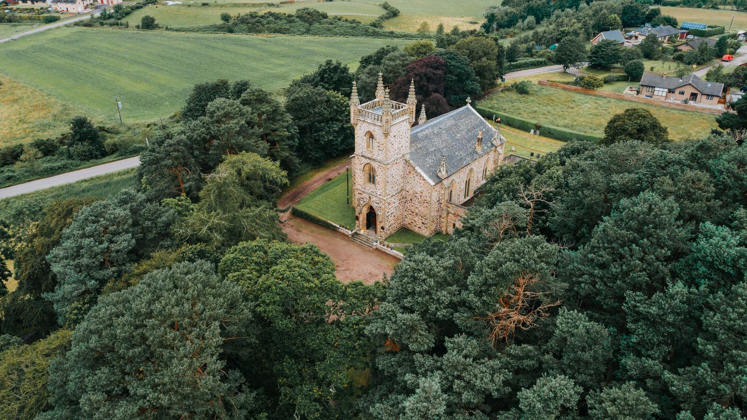 Aerial view of a historic stone church surrounded by lush green countryside and dense forest canopy.