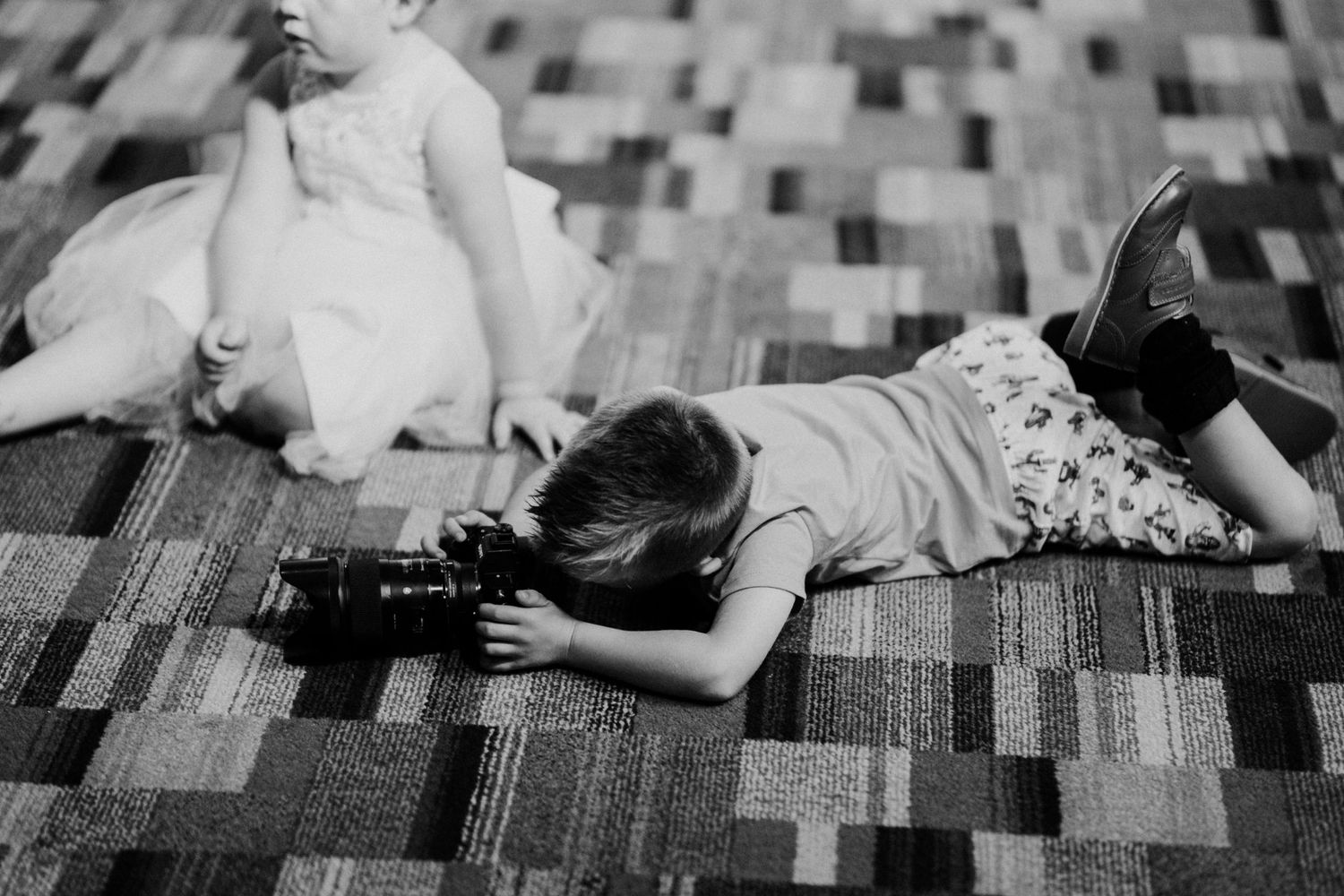 Black and white photo of a child lying on a patterned carpet, playing with a toy train.