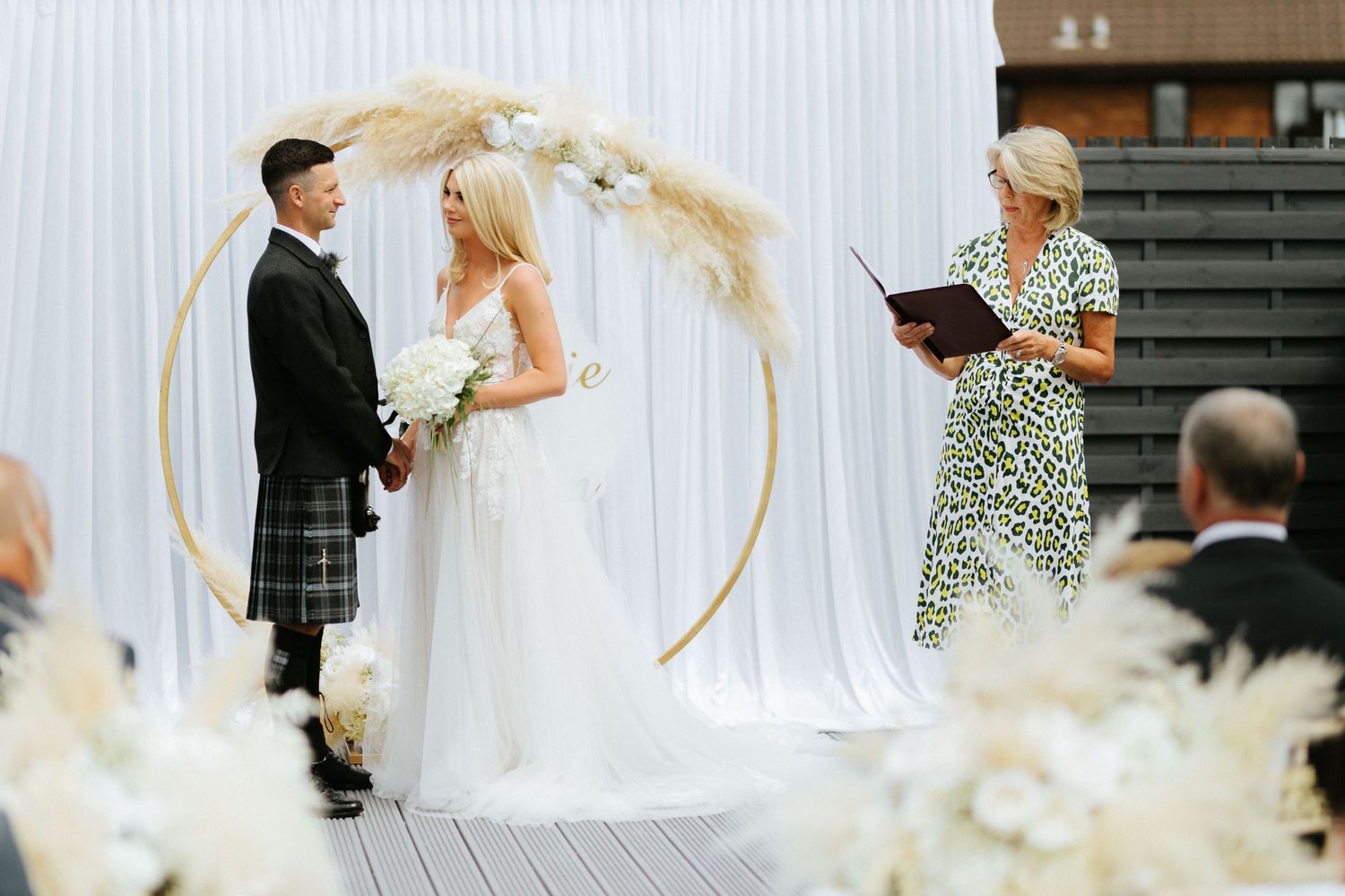 A couple stands under a circular floral arch during their outdoor wedding ceremony with pampas grass decor.