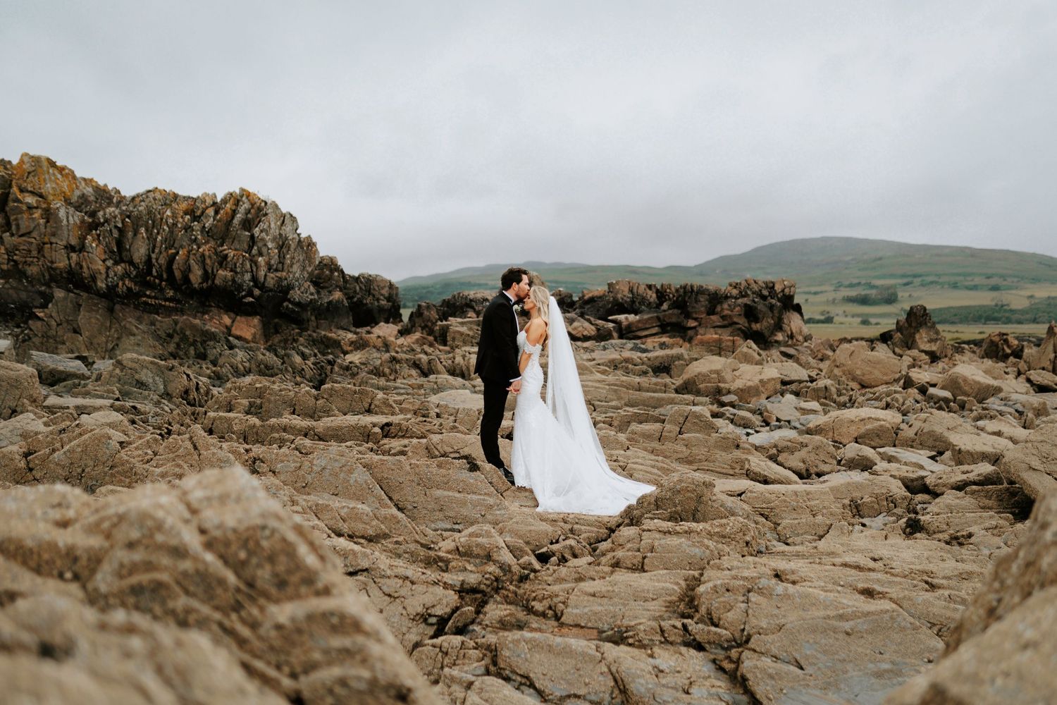 Two people in wedding attire embrace on rocky coastal terrain with rolling green hills in the background.