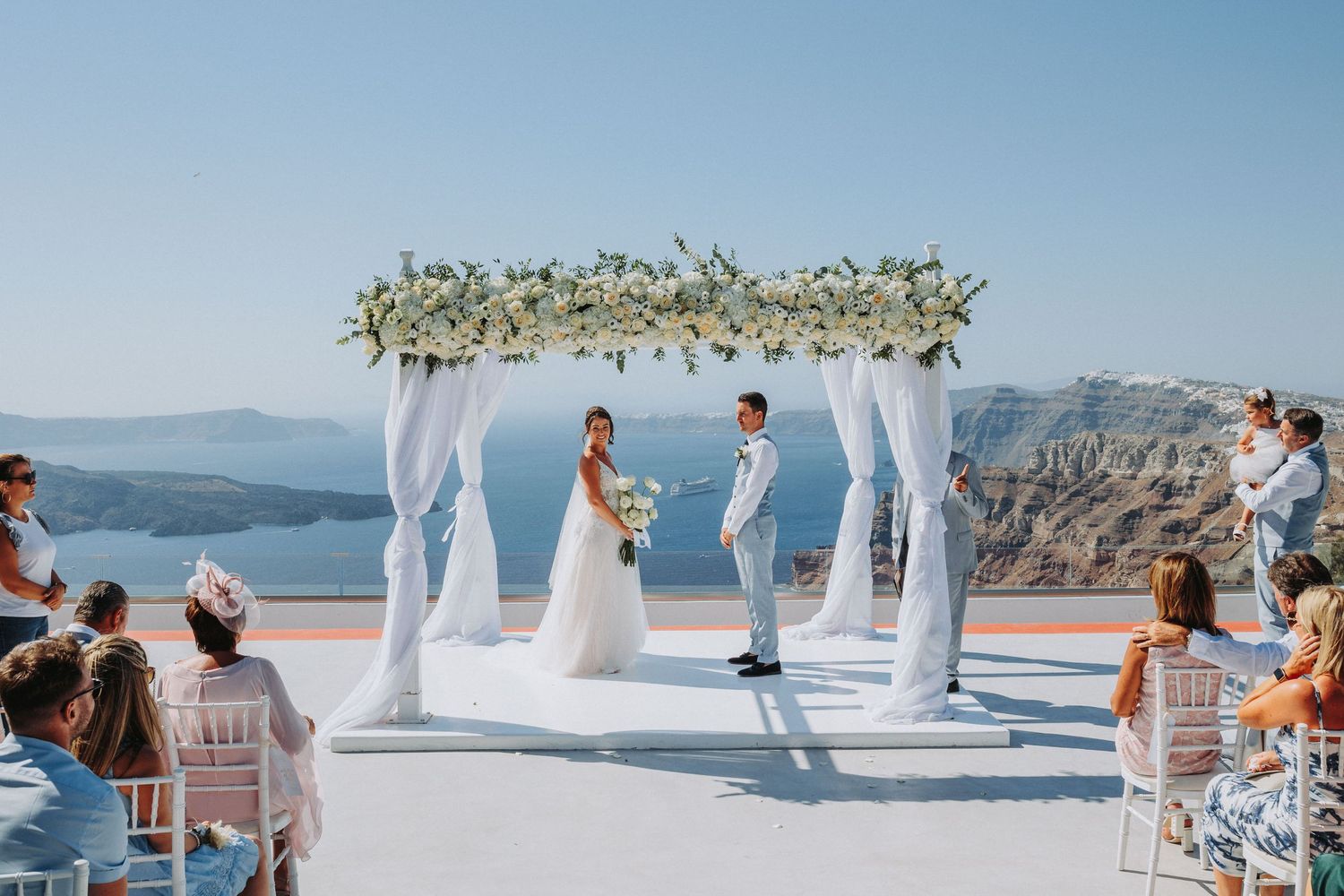 Romantic outdoor wedding ceremony overlooking the Mediterranean sea with white draped arch and floral decor in Santorini.