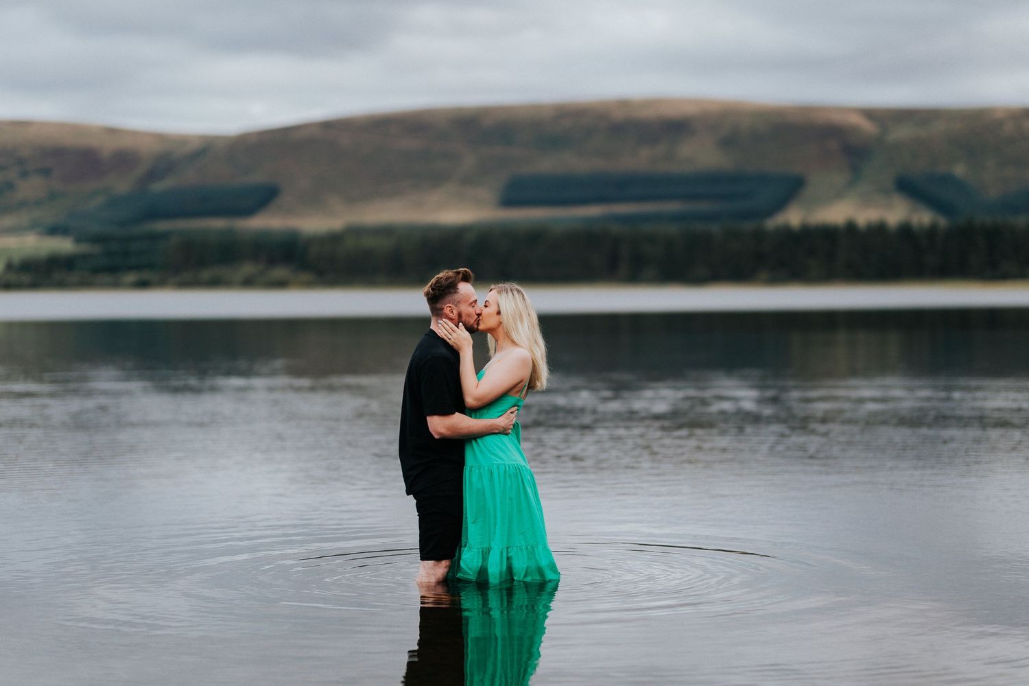 A couple embraces while standing in calm lake waters, with rolling hills in the background during twilight.