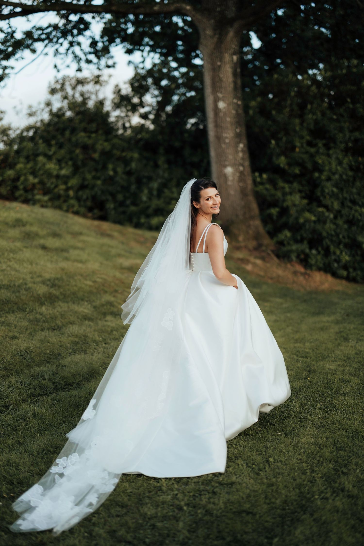 A dreamy bride in a flowing white wedding gown poses elegantly on a grassy hillside beneath tall trees.