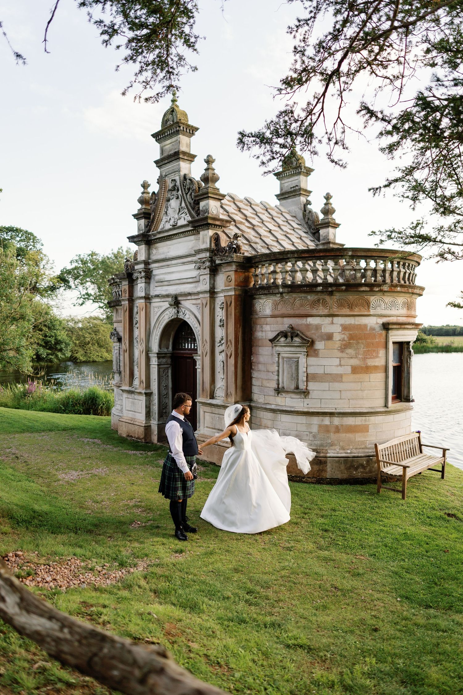 A romantic wedding photo sequence at a stone chapel with ornate architecture beside a lake on a grassy hillside.