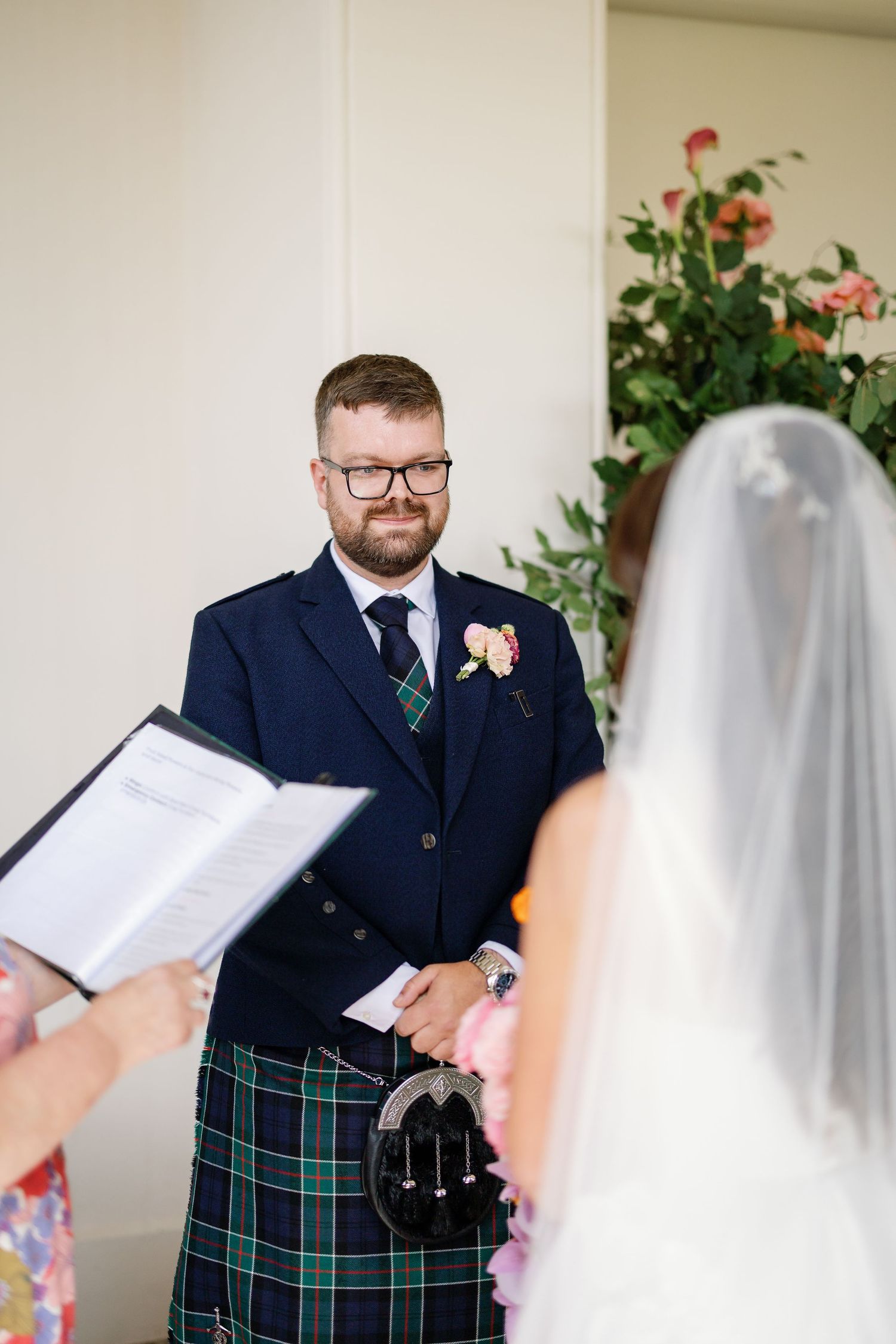 A wedding ceremony moment showing the groom in a navy suit with tartan kilt smiling during the vows.