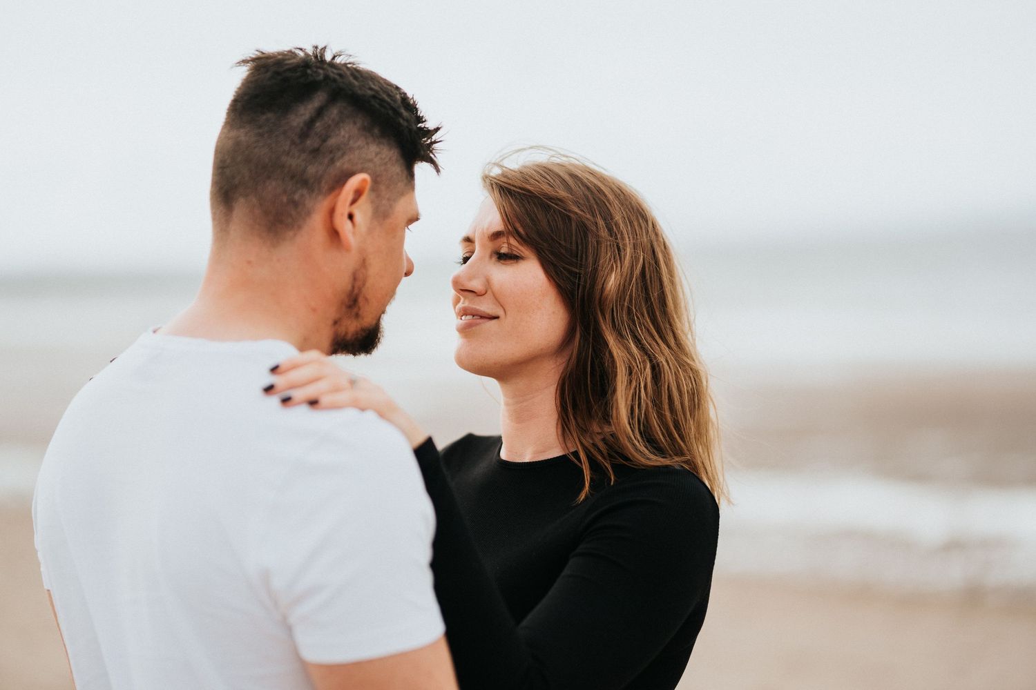 A couple shares an intimate moment together on a cloudy beach as they gaze at each other lovingly.