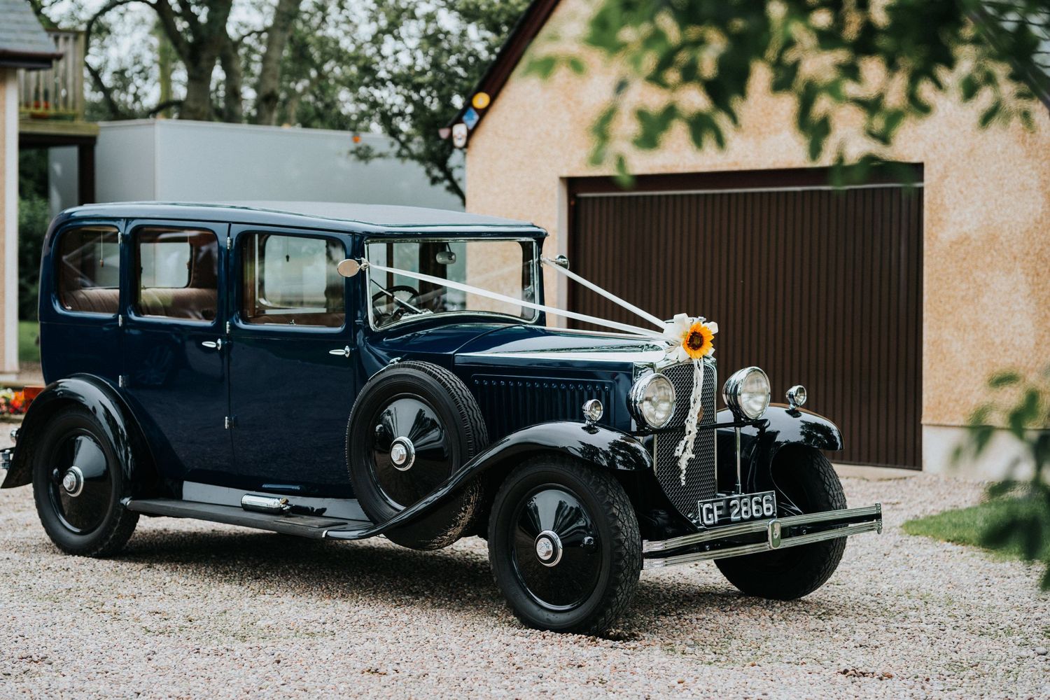 Vintage black 1930s luxury car parked on gravel driveway beside brown garage door and stone wall.