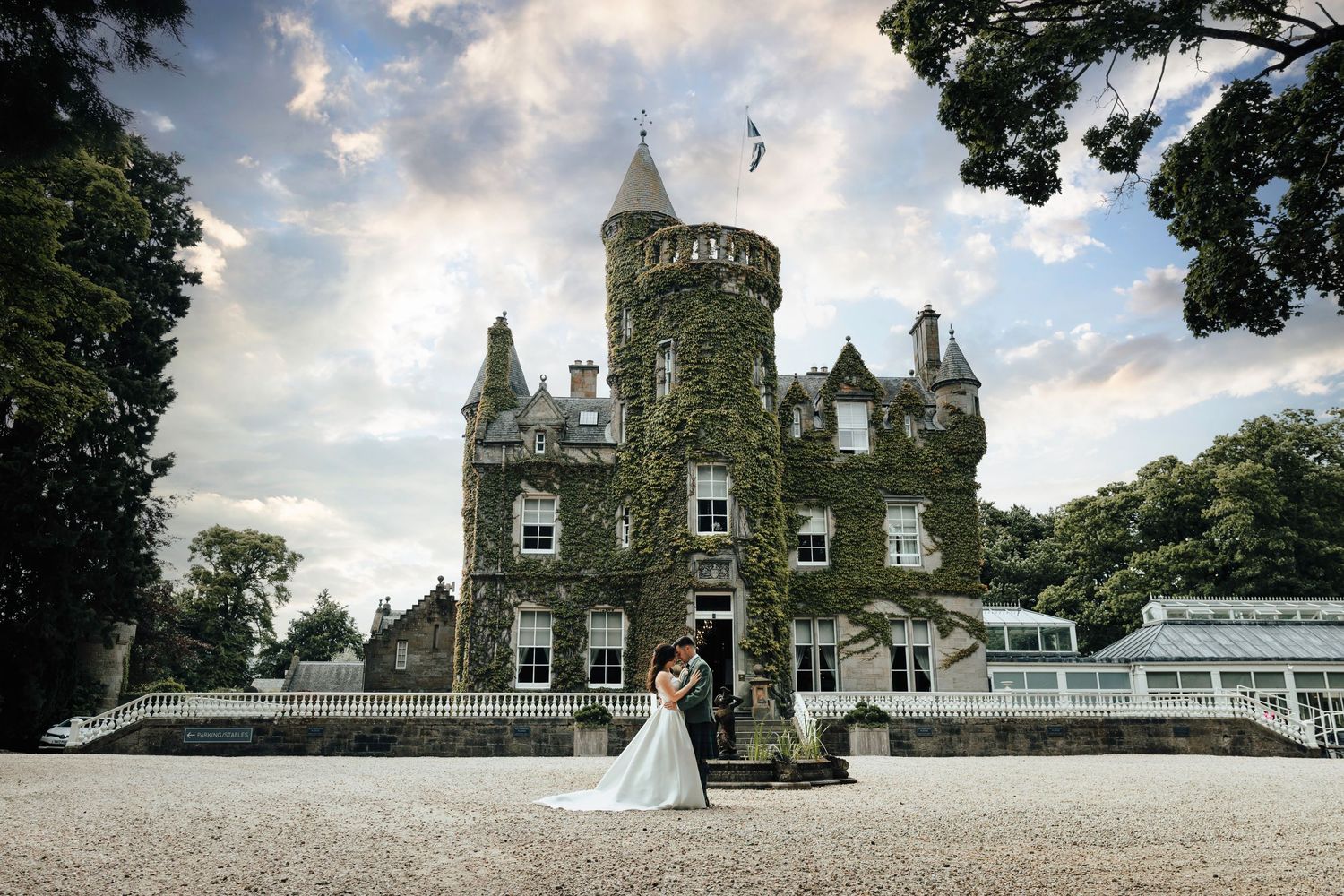 A romantic Victorian stone castle with tower features and a greenhouse extension stands against a cloudy sky. Carlowrie Castle