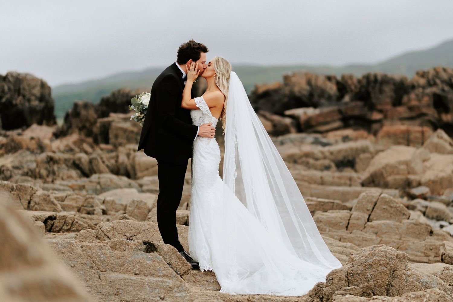 Wedding couple sharing an intimate moment on rocky terrain with mountains in the misty background.