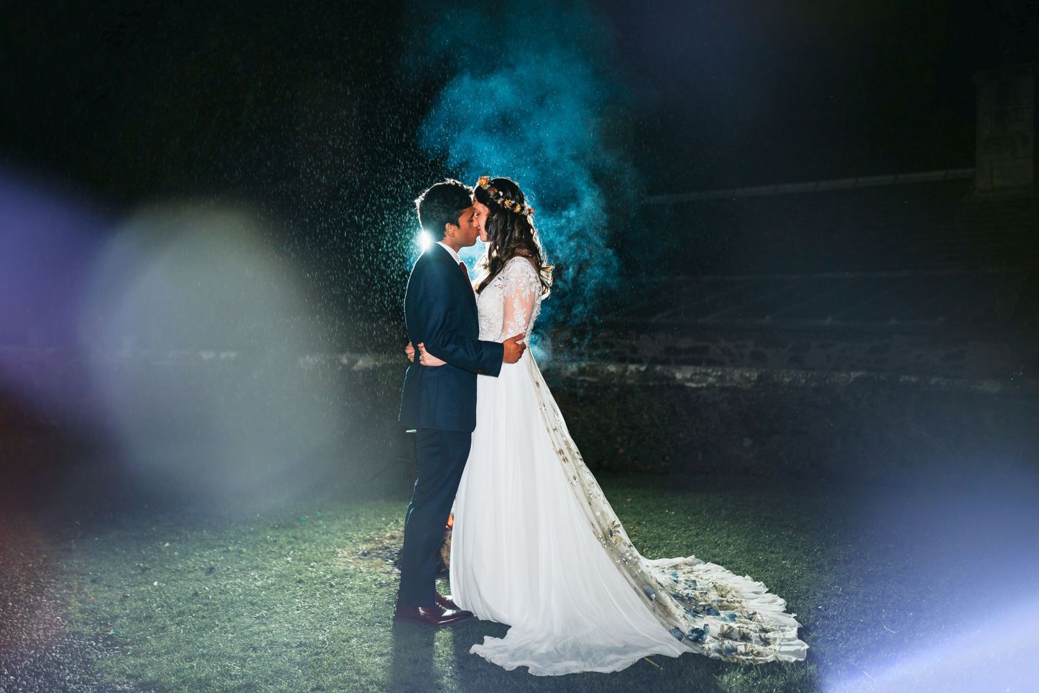A couple stands together in a dark garden with dramatic blue backlight and a flowing white wedding dress.