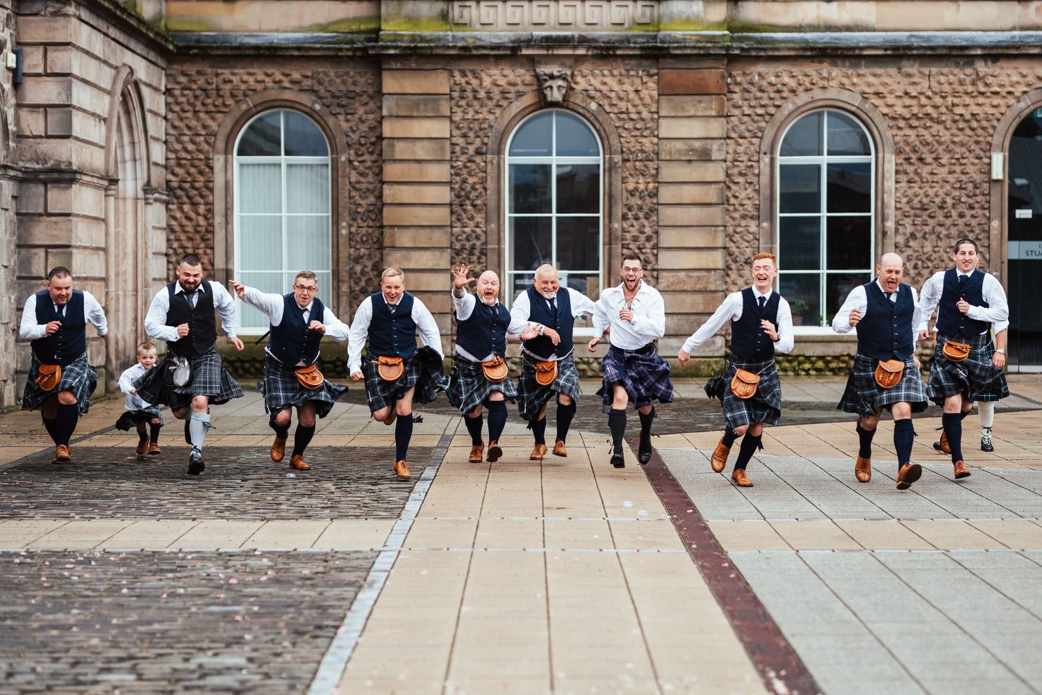 Groomsmen in coordinated attire kick their legs up while posing in front of an ornate brick building.