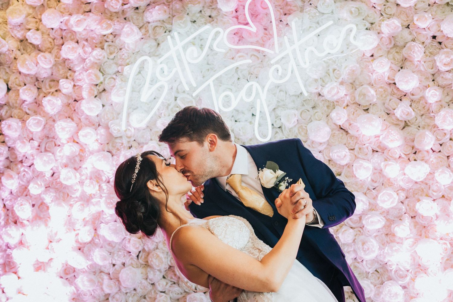 A couple dances together against a backdrop of pink floral decorations and neon signage at their wedding celebration.