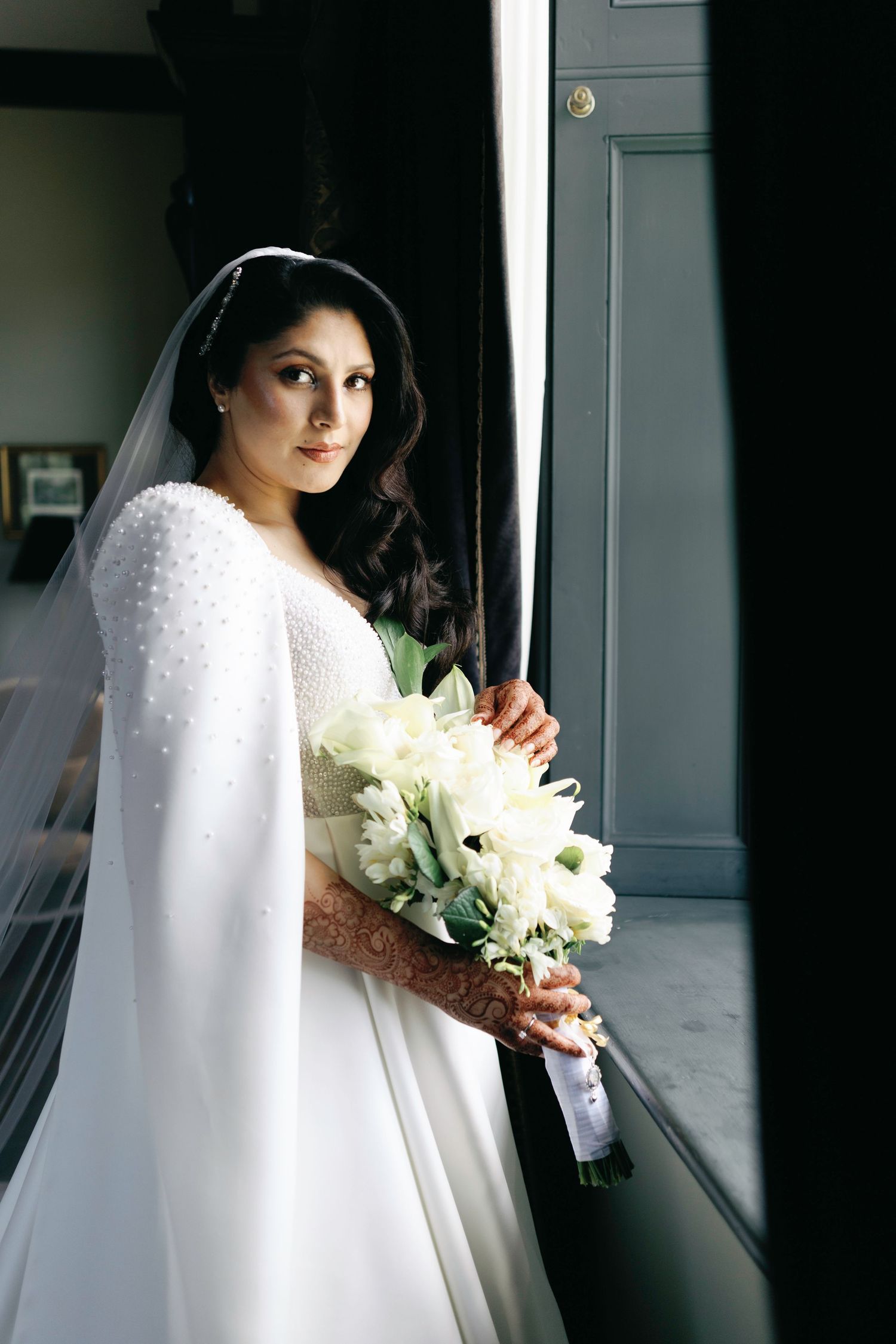 Beautiful bride in long white wedding dress and cape holding elegant white lily bouquet while standing by window.