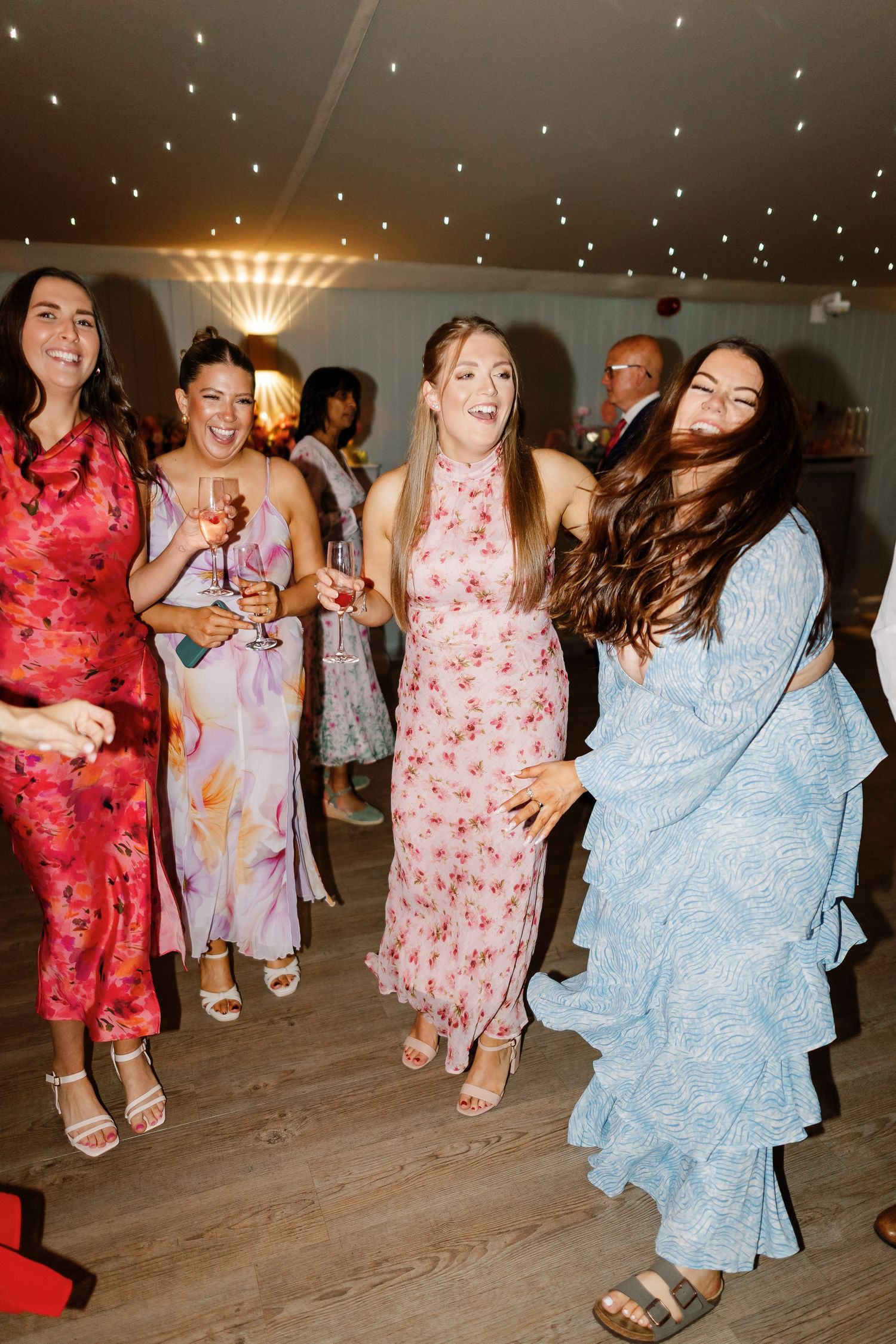 People dancing at a party wearing colorful floral and blue dresses on a wooden dance floor under twinkling lights.