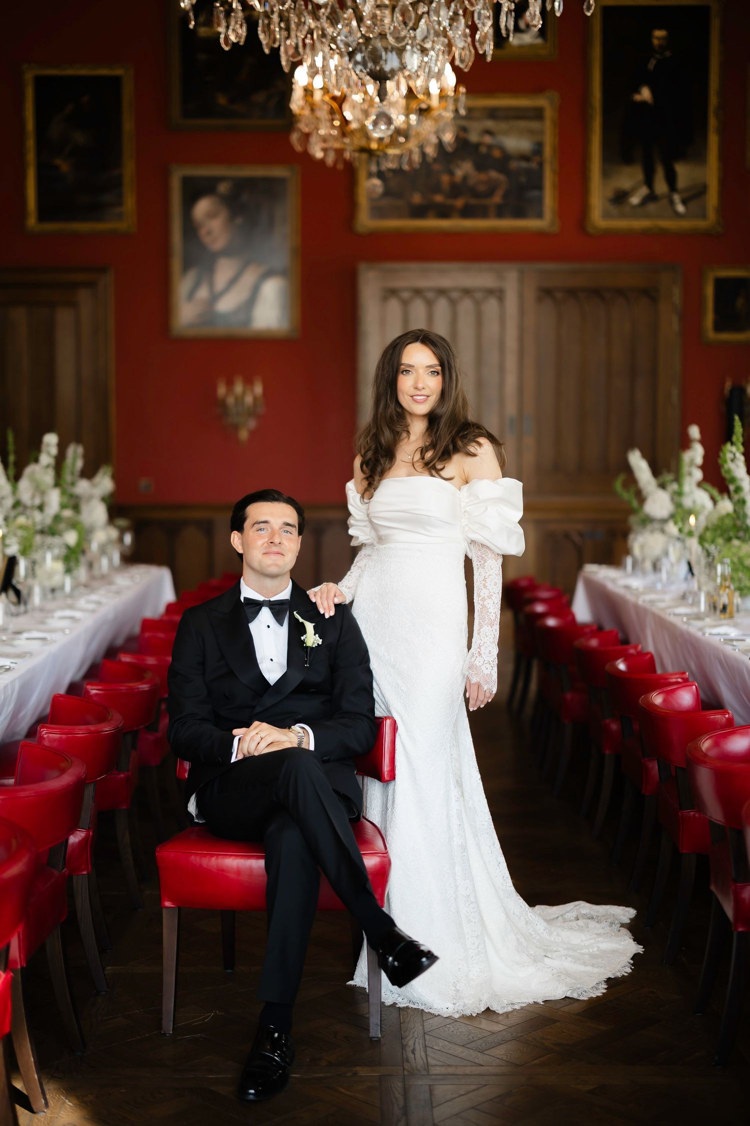 Elegant bride in off-shoulder white gown stands beside seated groom in tuxedo in ornate red wedding venue.
