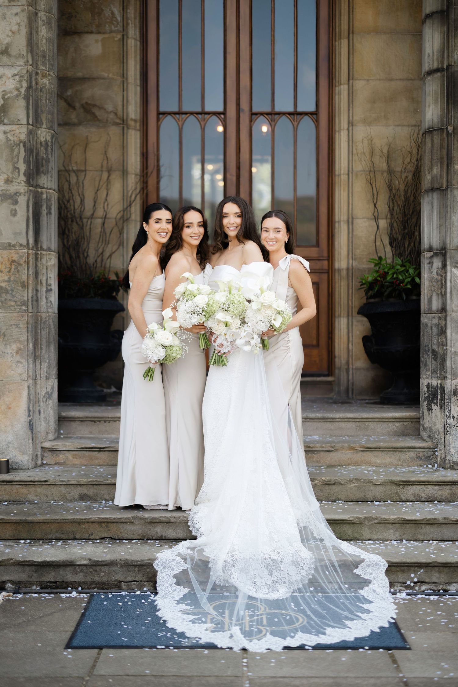 Bride in white lace gown with long veil poses with three bridesmaids in ivory dresses holding white bouquets.