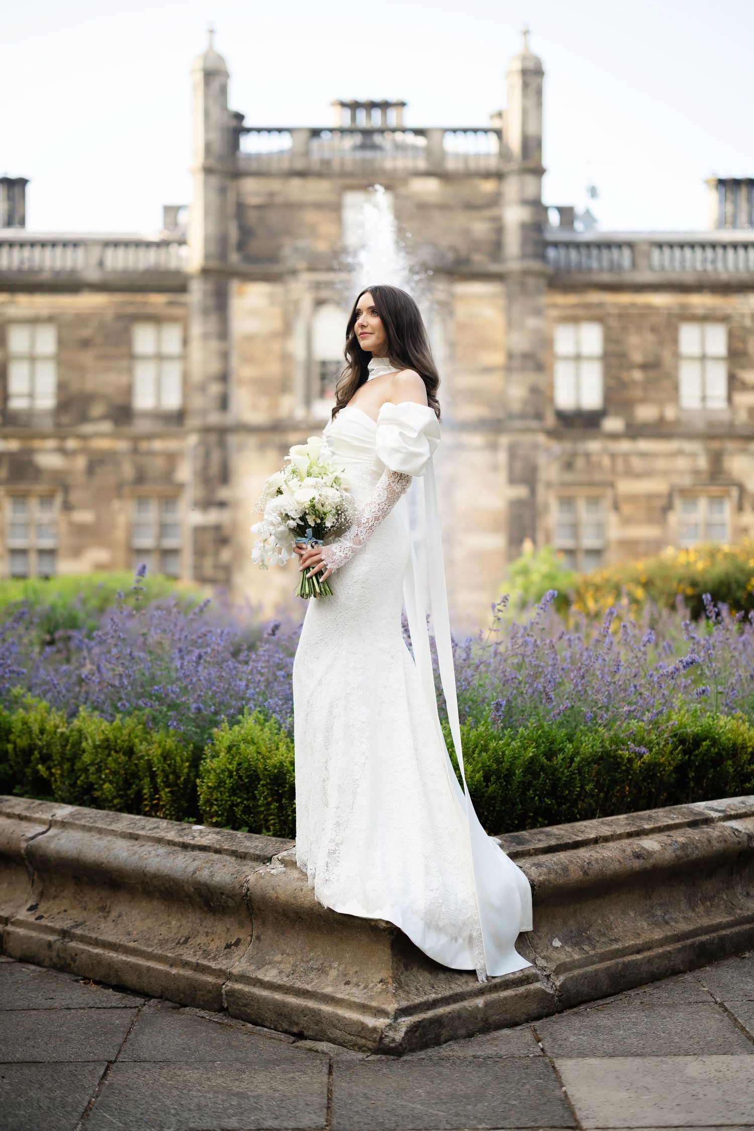 Bride in white off-shoulder gown holds bouquet before historic stone mansion with lavender gardens.
