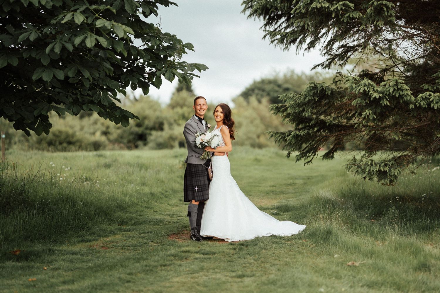 A couple stands together on a lush green lawn framed by trees during their outdoor wedding celebration.