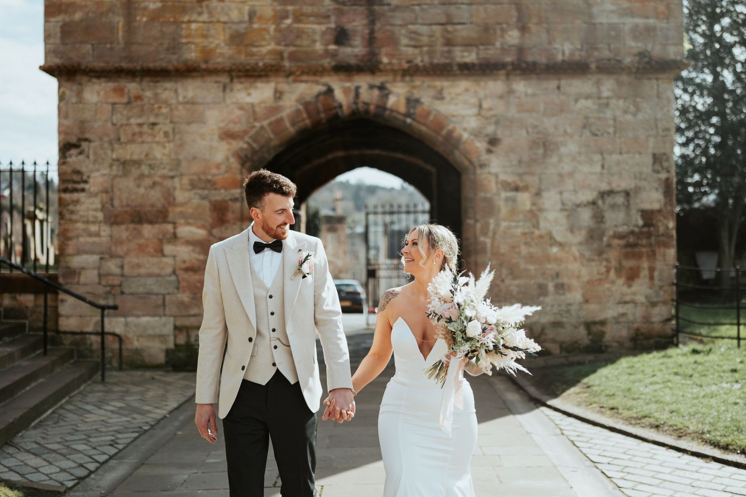 A newlywed couple walking through a stone archway at a historic venue on their wedding day.