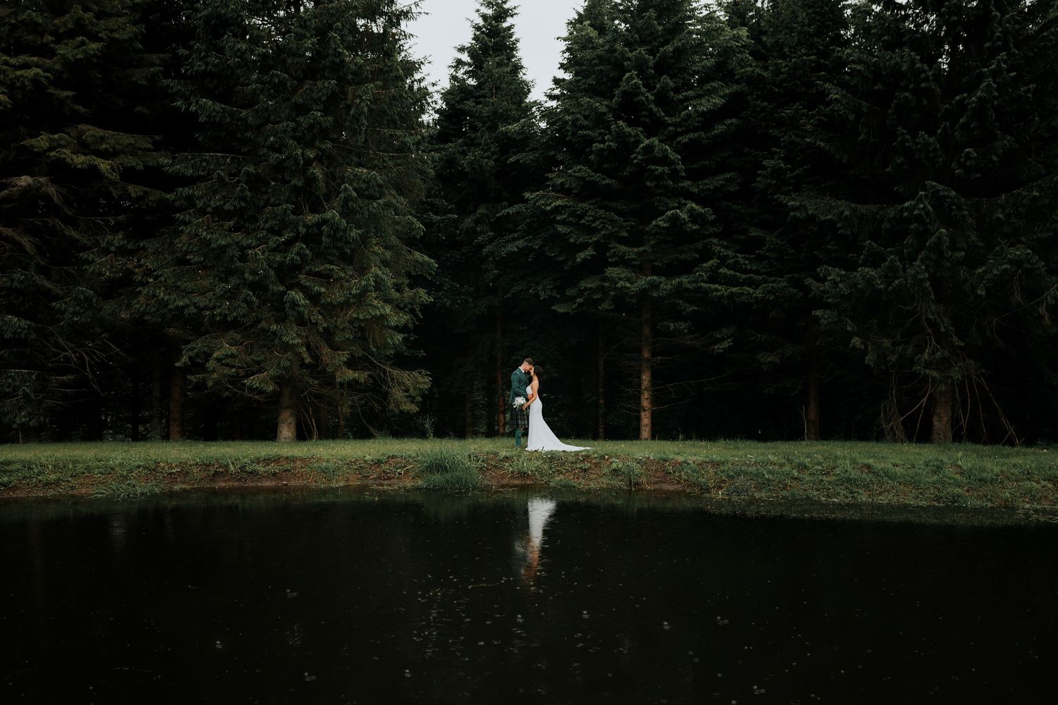 A couple stands at the edge of a serene pond surrounded by evergreen trees, their reflection mirrored in the still water.