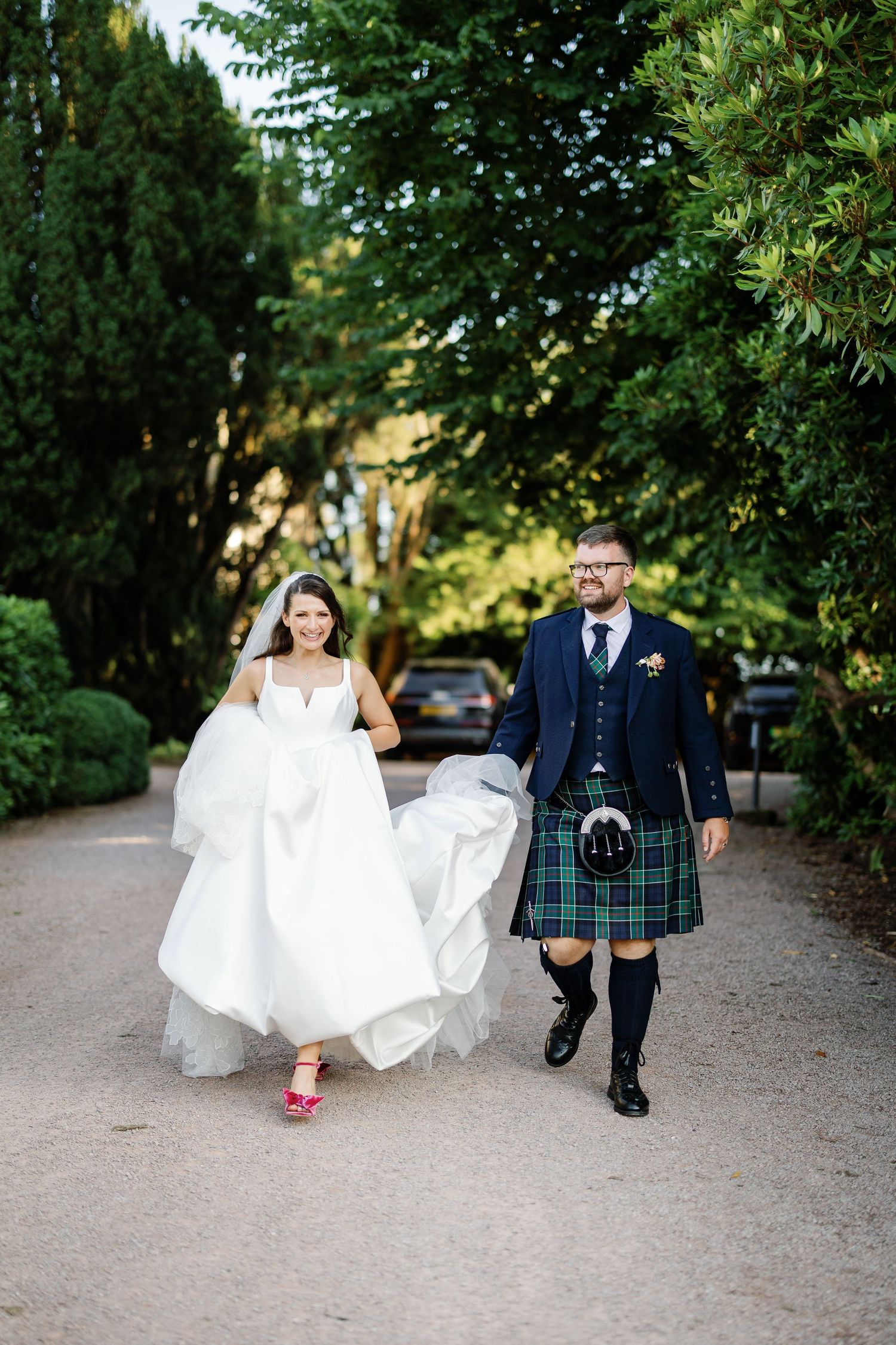 A wedding couple walks together on a garden path, with the groom wearing a traditional Scottish kilt and the bride in white.