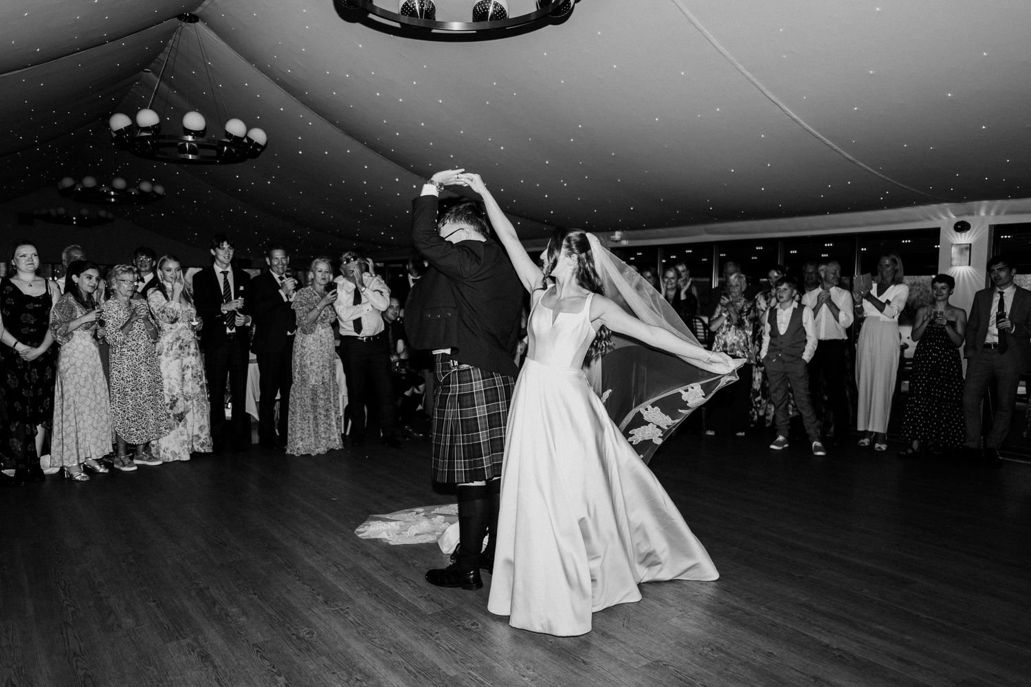 Wedding guests gather on dance floor during reception celebration with string lights overhead.
