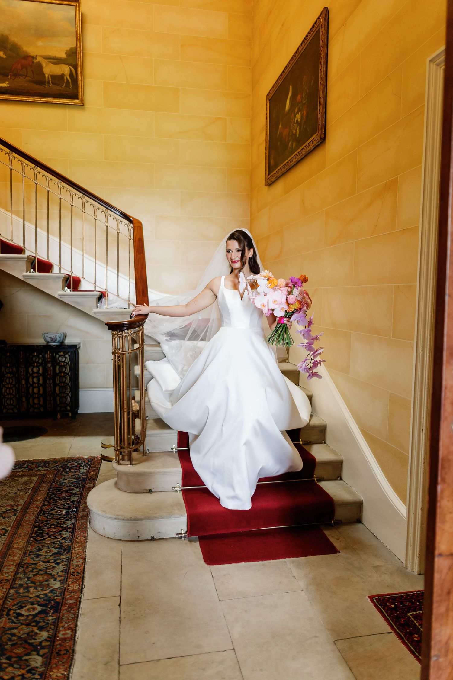 A bride in a flowing white wedding dress twirls on an elegant staircase with red carpet and ornate railings.