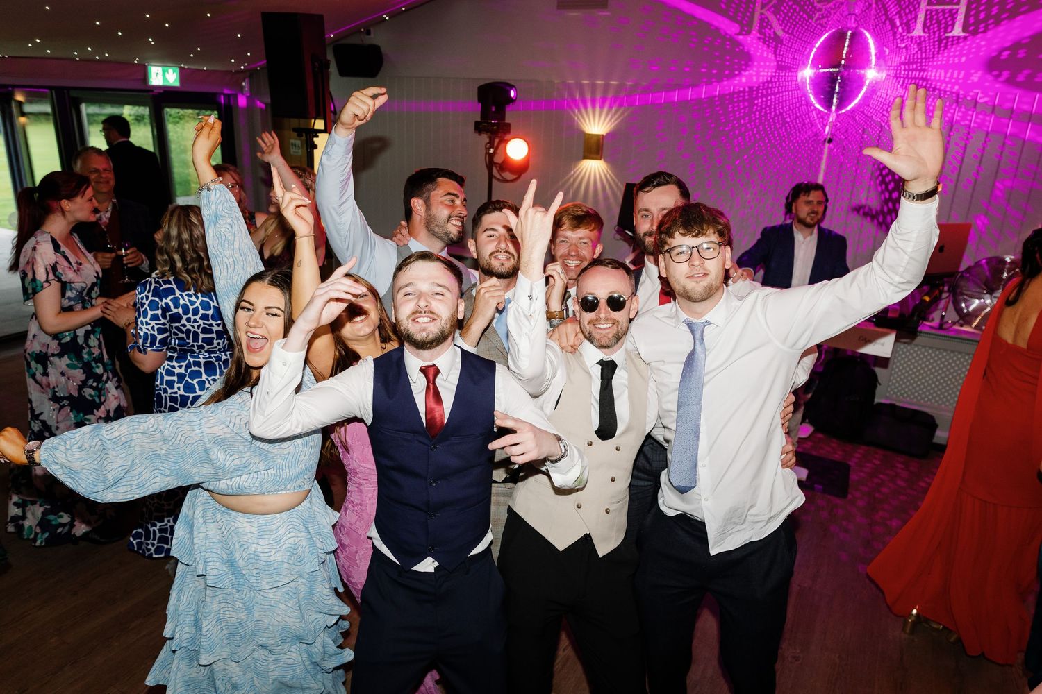 Wedding guests dancing and celebrating on the dance floor at a reception with pink lighting and festive atmosphere.