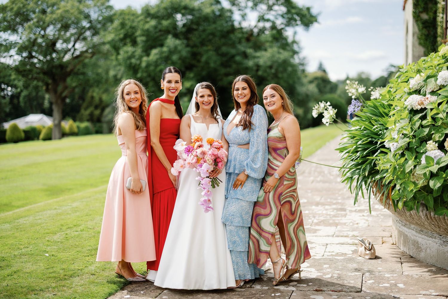 A group of people in pastel and coral colored dresses stand together outdoors on a sunny day at a formal event.