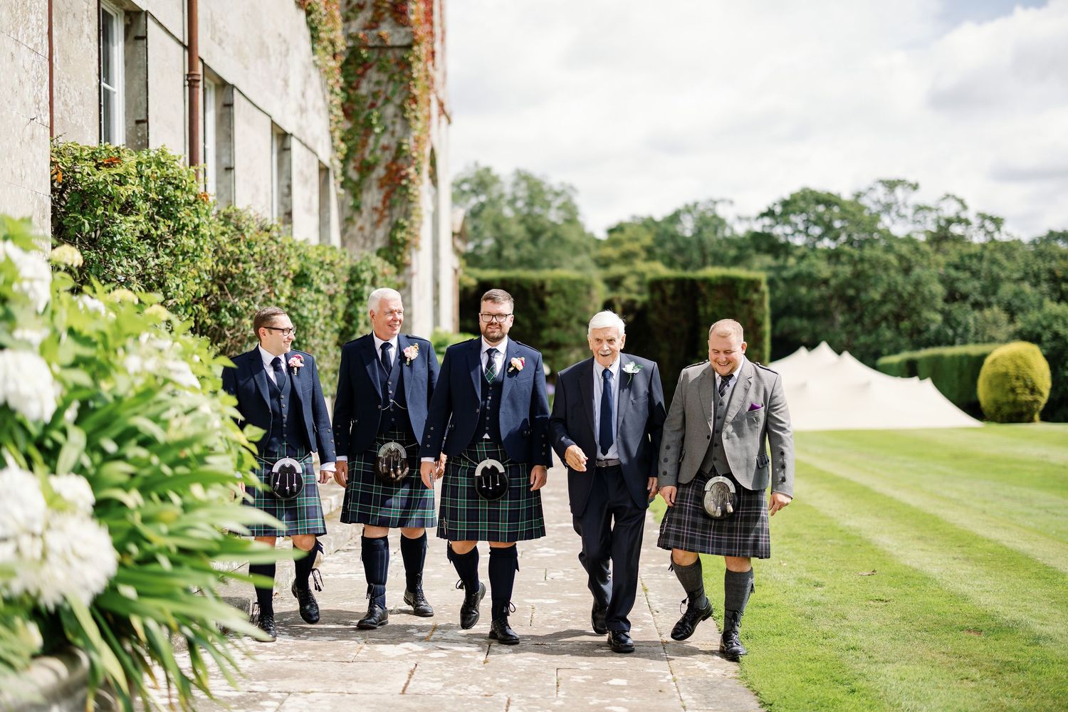 A group in traditional Scottish kilts walking along a stone path beside a manicured garden and lawn.